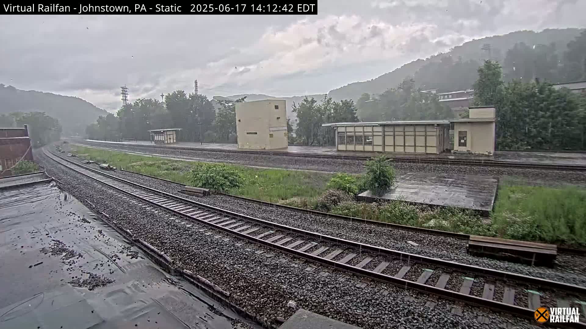 Rain falls on a railway station with multiple tracks and small buildings nestled in a valley surrounded by lush green hills.