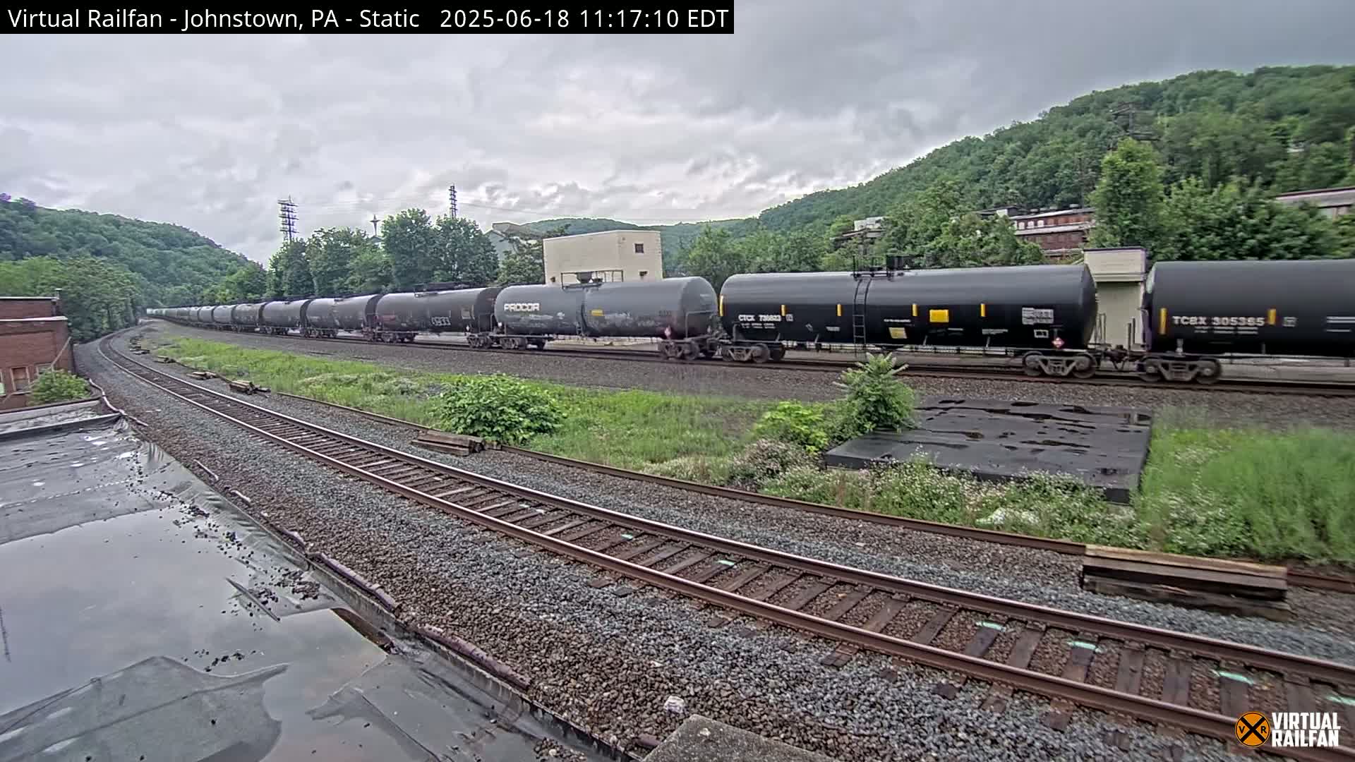A long freight train of black tanker cars sits on a railway track beneath an overcast sky, surrounded by green hills and vegetation.