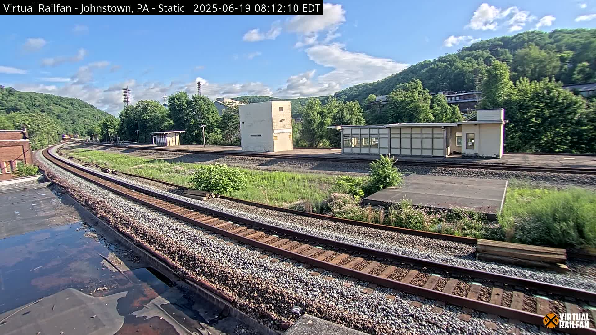 A train track curves through a grassy area, past several small buildings and a hill covered in green trees under a mostly sunny sky.