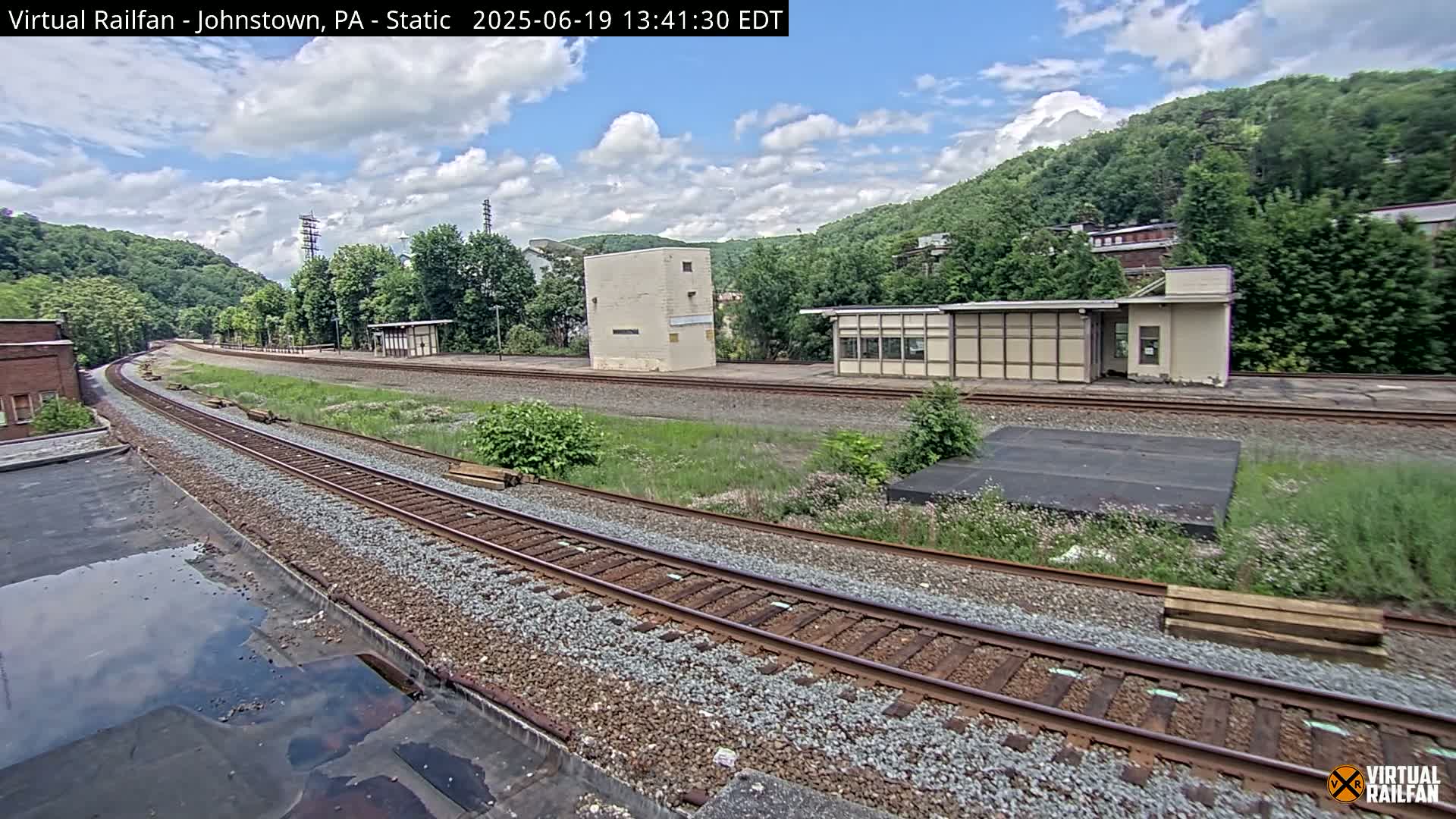 A partly cloudy day reveals railroad tracks curving through a grassy area with small buildings and a hillside covered in green trees in the background.