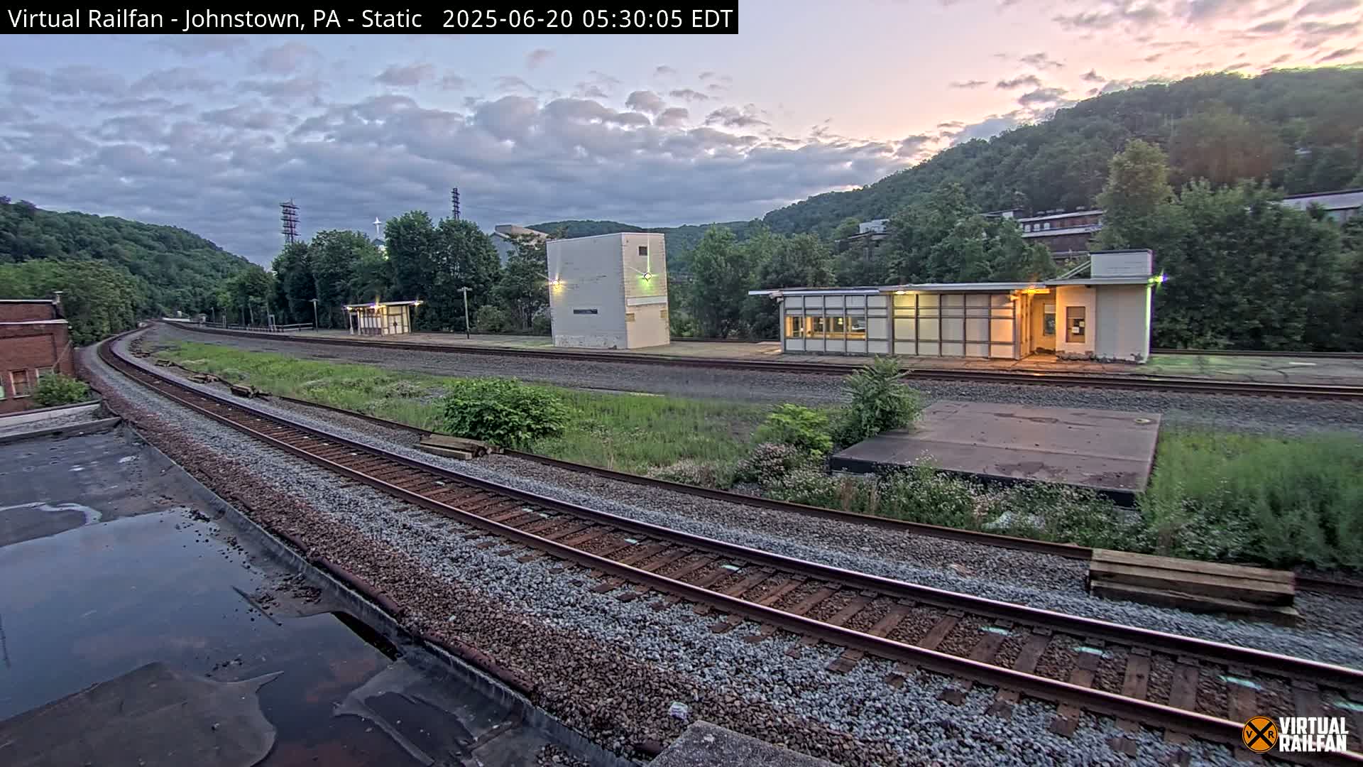 A train track curves through a grassy area with several small buildings near a hillside at dusk.