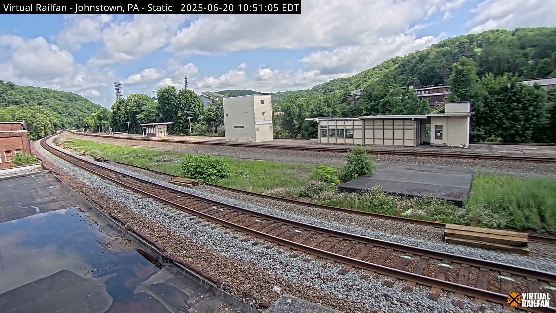 A partly cloudy day reveals a railway scene with two train platforms and a small building, nestled amongst overgrown vegetation and hills.