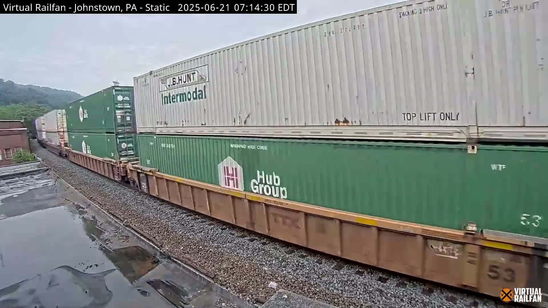 A long freight train carrying multiple shipping containers moves along railway tracks, past a building and a hillside, under overcast skies.