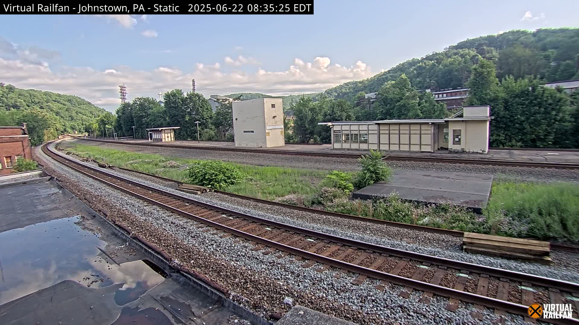 A mostly sunny view of train tracks curving gently through a grassy area with some low buildings and green hills in the background.
