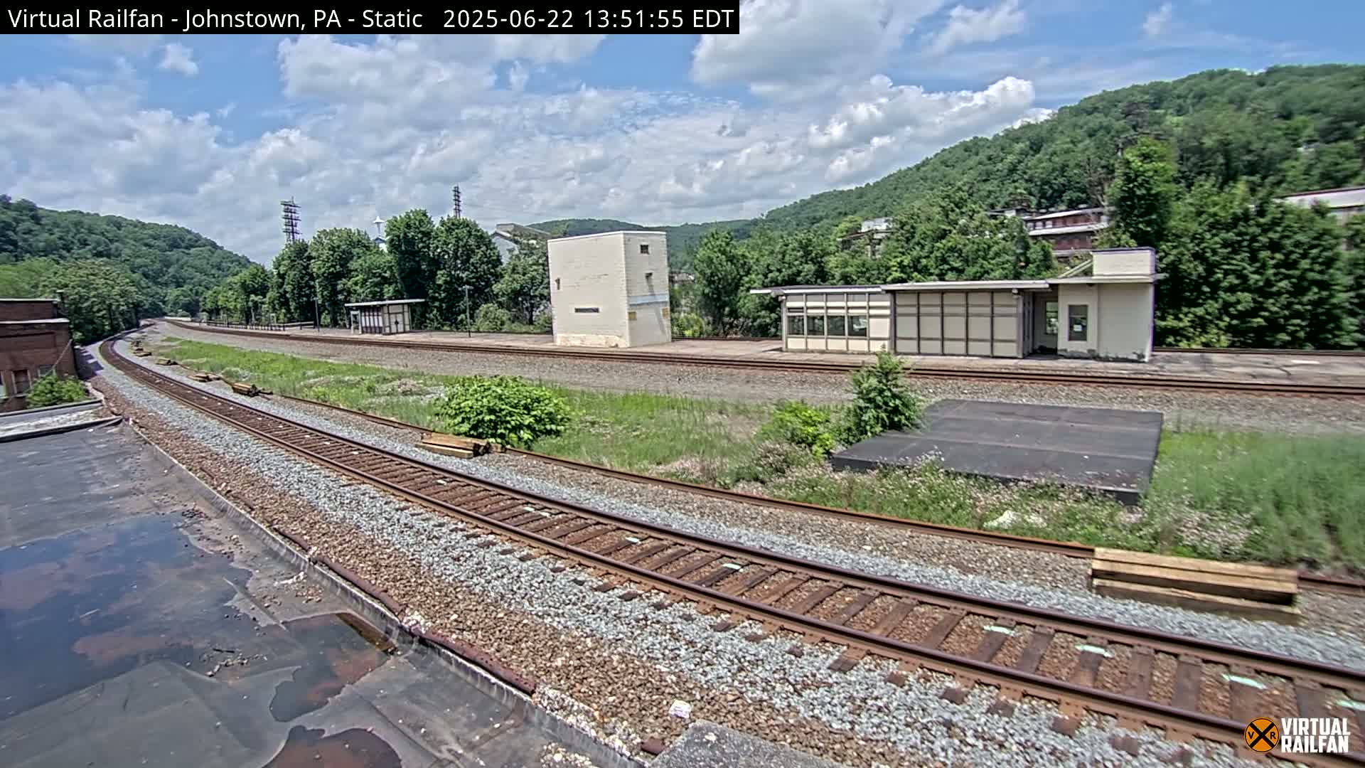 A sunny day view of train tracks curving through a grassy area, with small buildings and a hillside covered in green trees in the background.