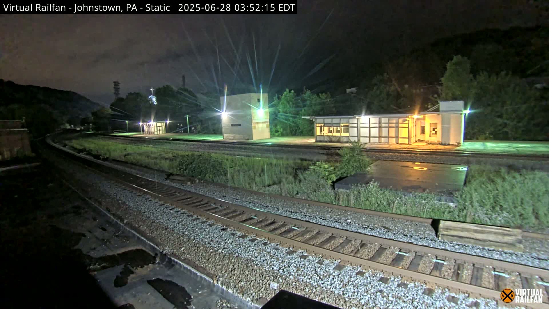 At night, a train station with several small buildings sits beside a curving set of train tracks against a backdrop of dark hills.