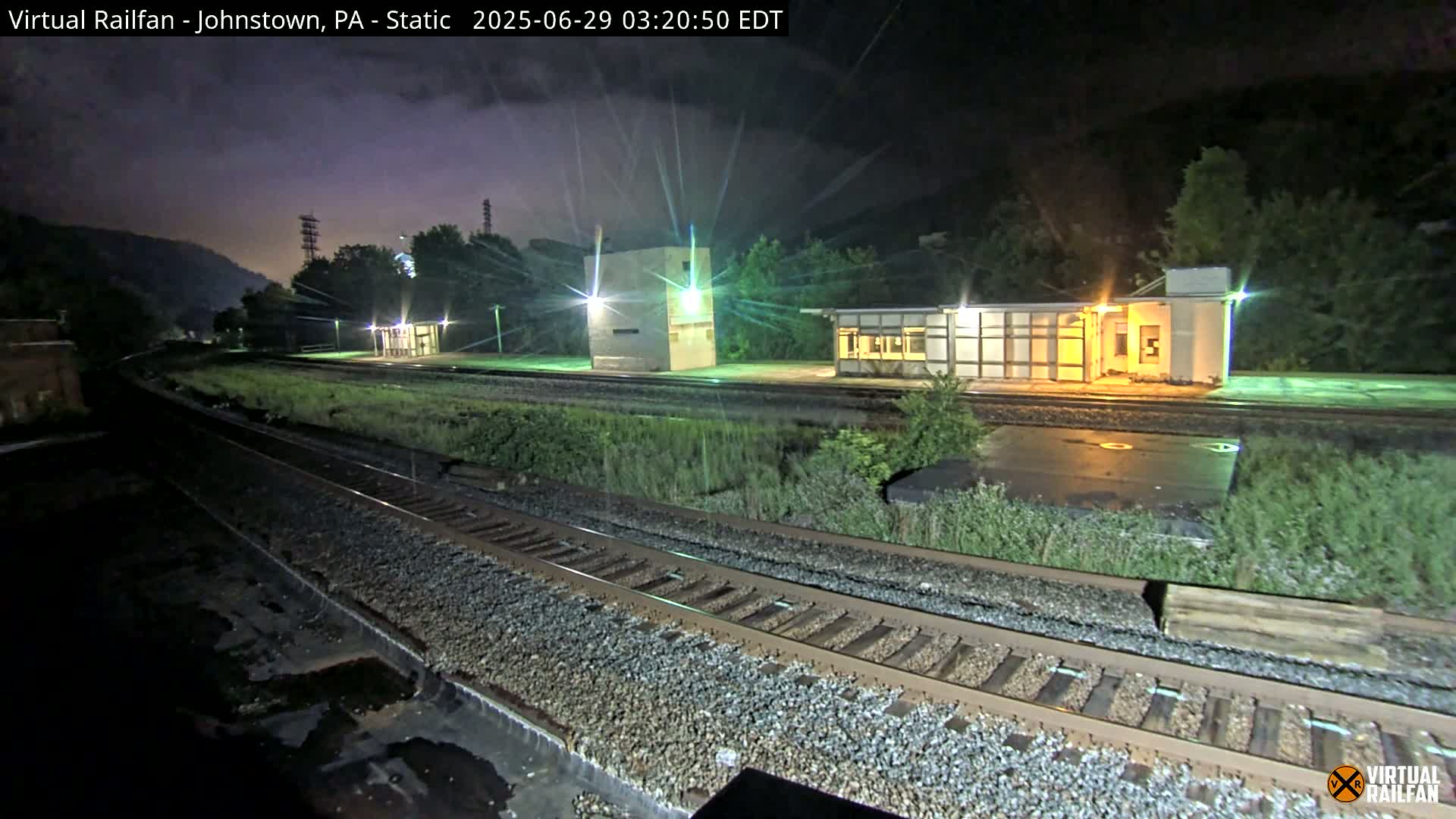 A nighttime view of a train station platform with several buildings and train tracks, under a dark sky.