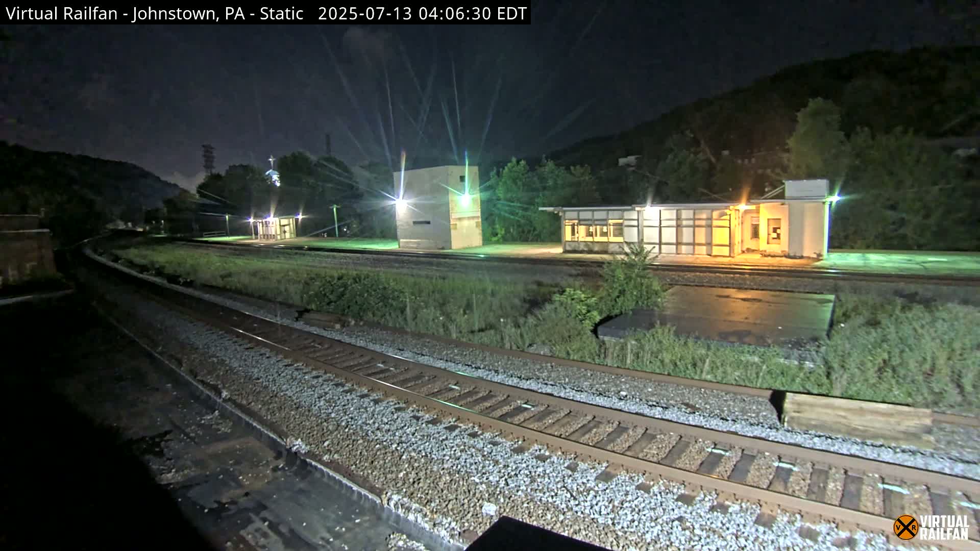 A nighttime view of a train station platform and railway tracks, illuminated by artificial lights, with hills in the background.