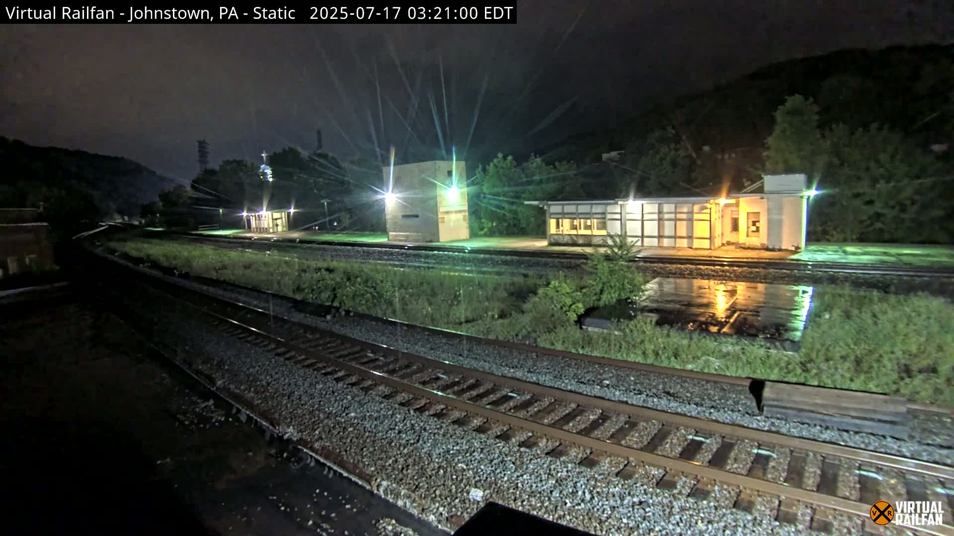A nighttime view of a rain-slicked train station platform and tracks, with several buildings illuminated under a dark sky.