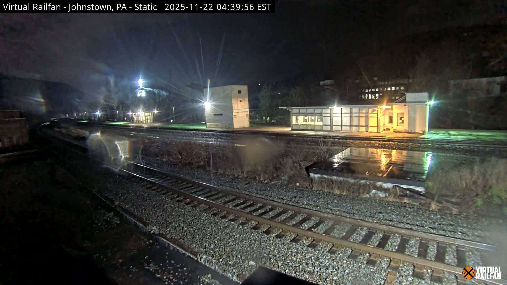 A nighttime scene depicts multiple wet railroad tracks curving into the distance alongside industrial-style buildings, all illuminated by artificial lights under rainy conditions.
