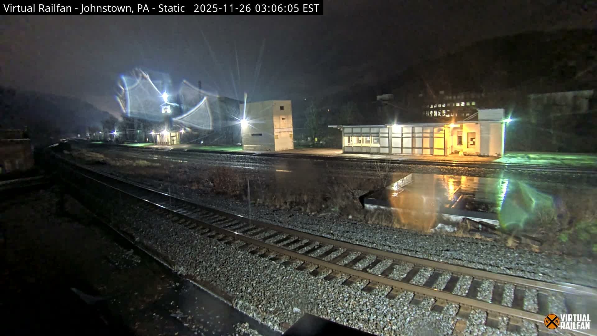 Railroad tracks stretch across wet ground in a nighttime scene, flanked by illuminated buildings and dark hills under what appears to be light rain or mist.