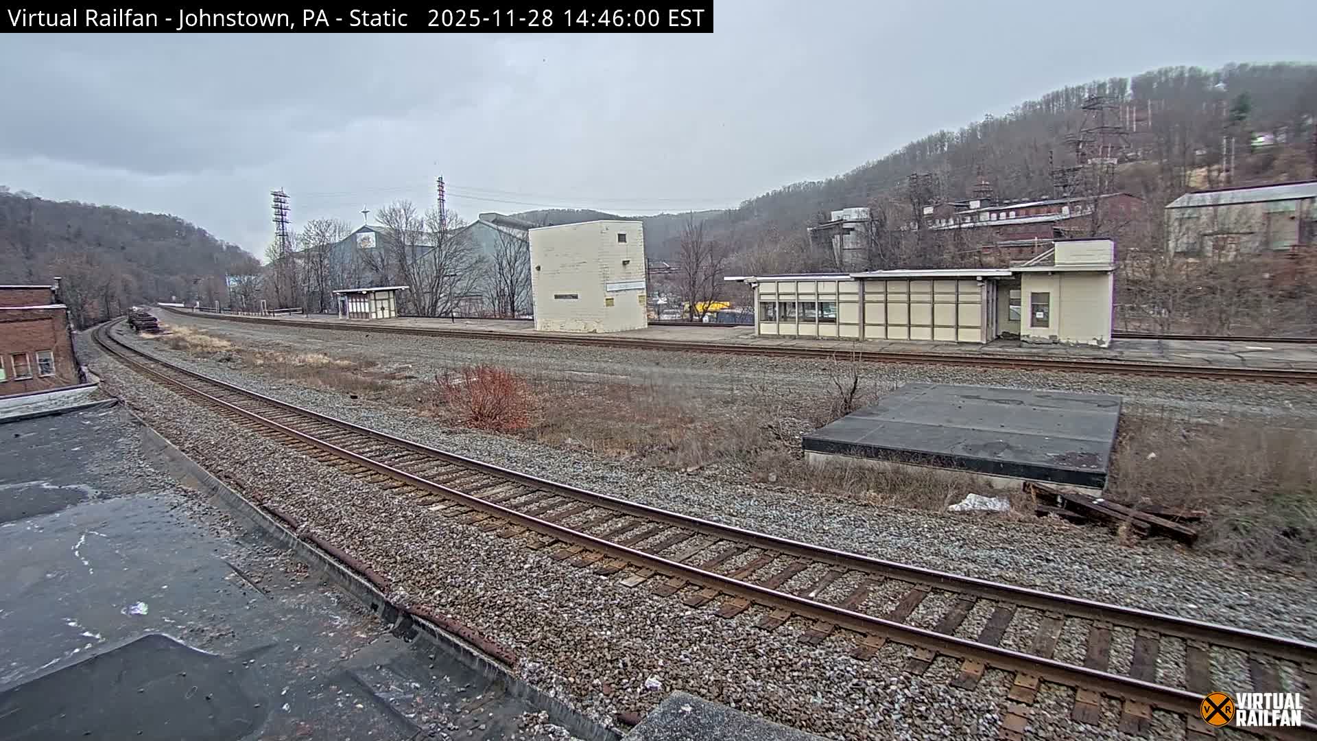 Multiple railroad tracks curve through an industrial valley bordered by bare hills and various buildings, including what appears to be a disused station, all under an overcast sky.