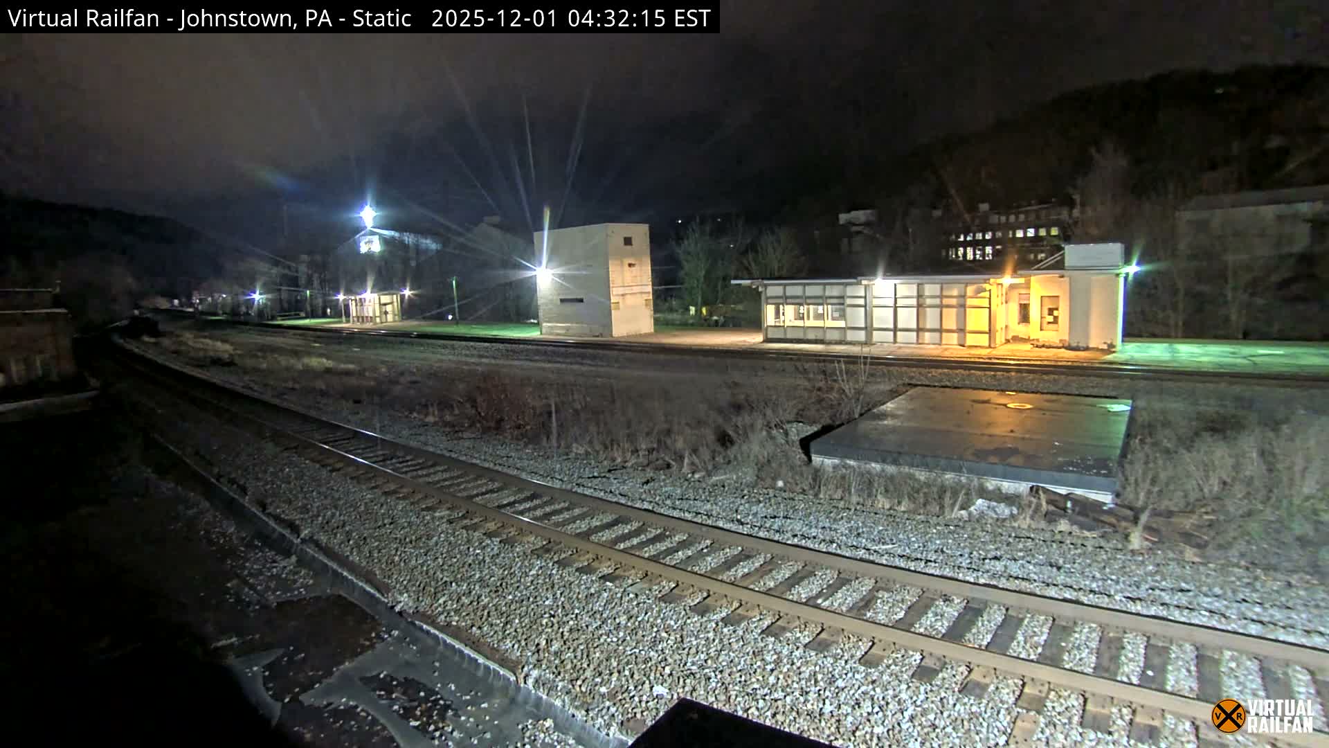 A dark night view reveals multiple railway tracks alongside illuminated industrial buildings and bare vegetation, set against distant hills under a clear, dry sky.