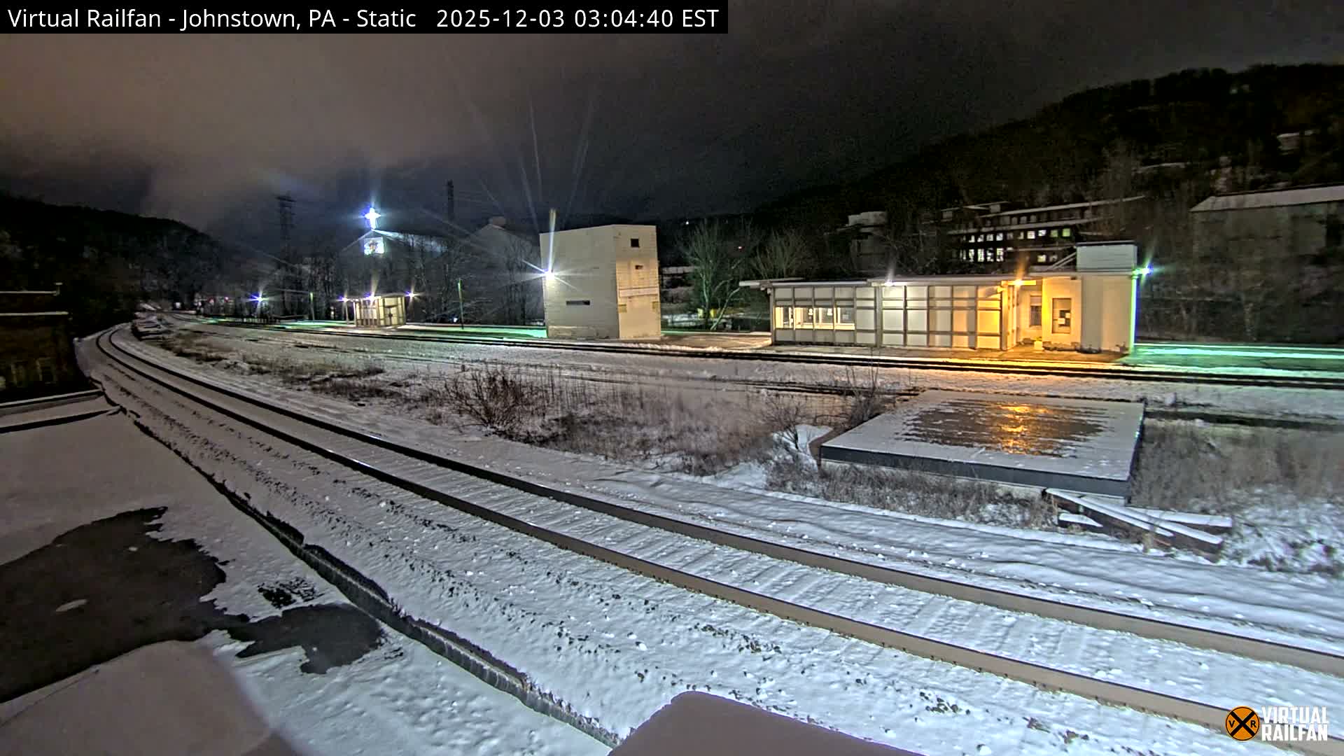 At night, a snow-covered rail yard features multiple tracks leading past illuminated industrial buildings and a station, set against dark, tree-covered hills under a cloudy sky.