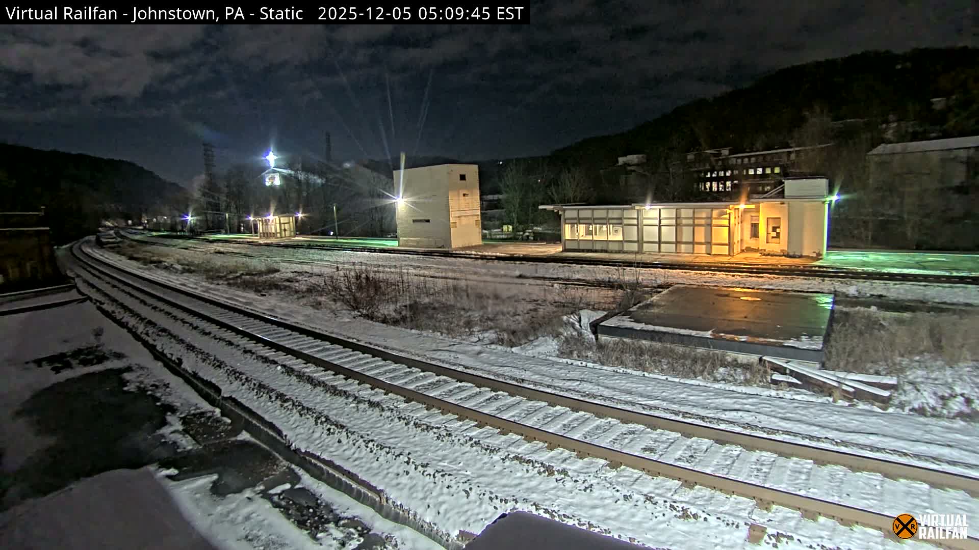 A snowy railroad yard is seen at night, featuring multiple tracks and illuminated industrial-style buildings nestled in a valley under a cloudy sky, with light snow covering the ground.