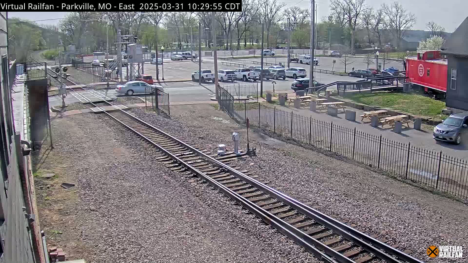 A sunny day shows railroad tracks intersecting a road near a parking lot with several cars, picnic tables, and a red caboose.