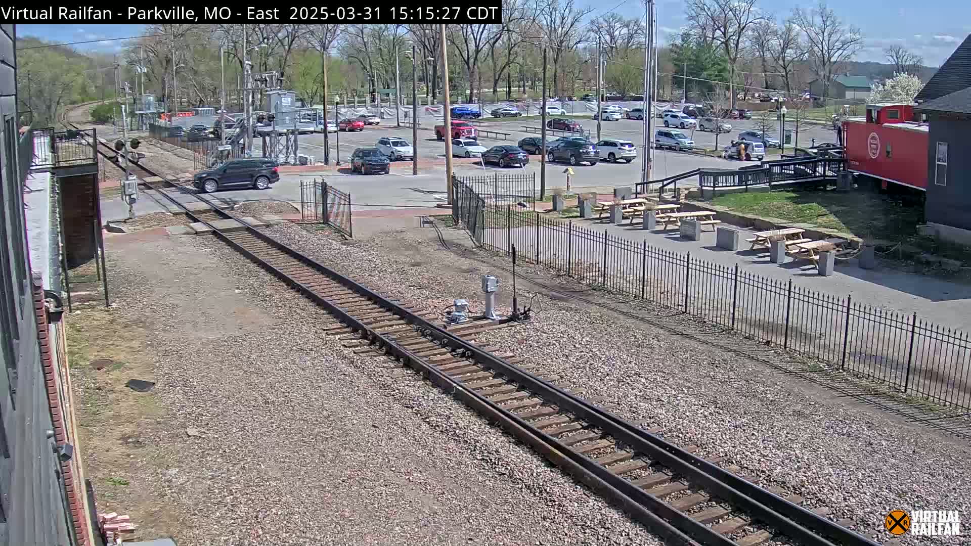 A sunny day shows a parking lot, a red caboose, picnic tables, and railroad tracks intersecting with a road.