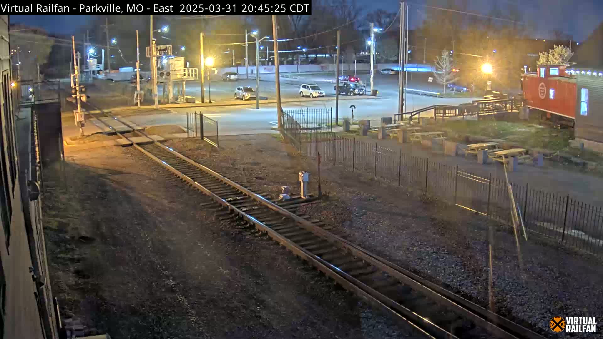At night, railroad tracks intersect with a road near a parking lot and a building that resembles a converted train car, with several cars visible in the parking lot.