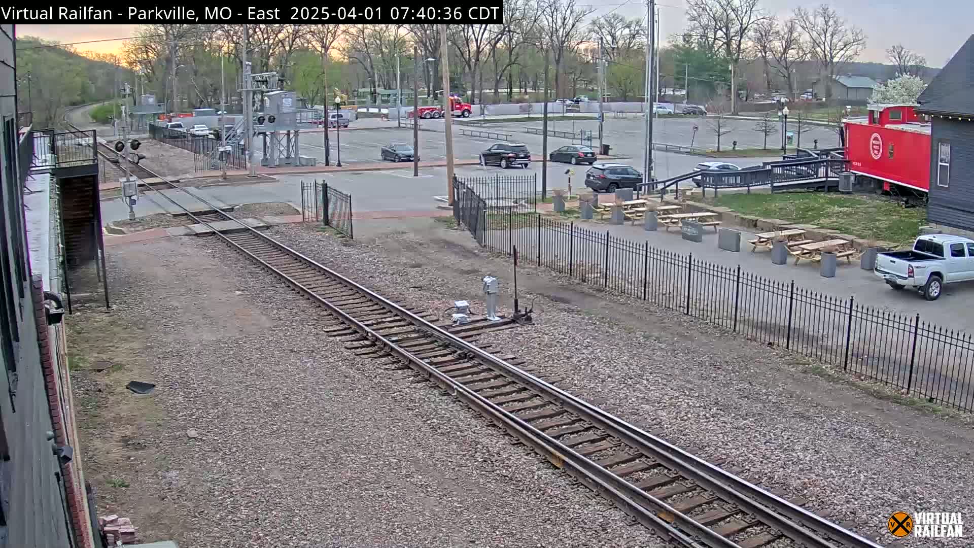 Railroad tracks run alongside a parking lot with several cars and a red caboose, all under a partly cloudy sky.