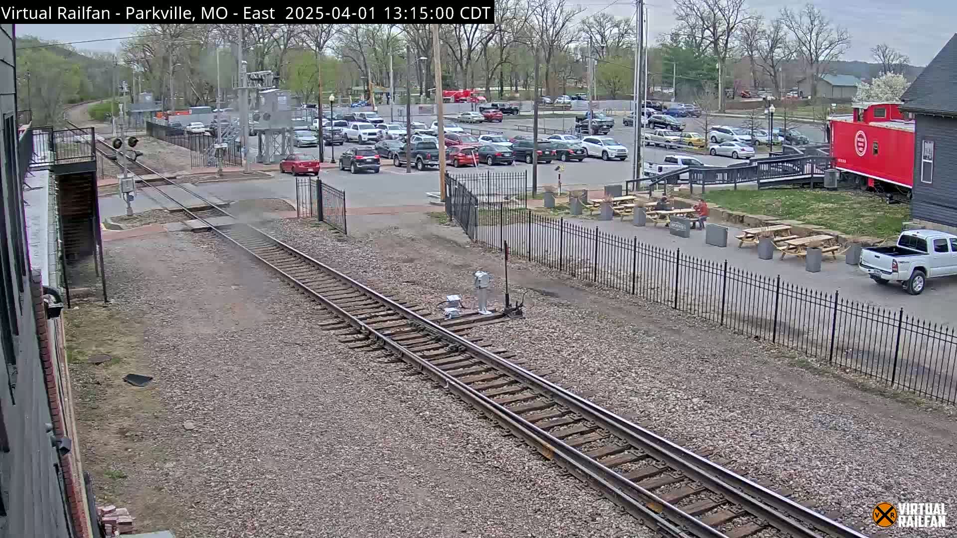 A railroad crossing with a parking lot full of cars, picnic tables, a red caboose, and a partly cloudy sky.