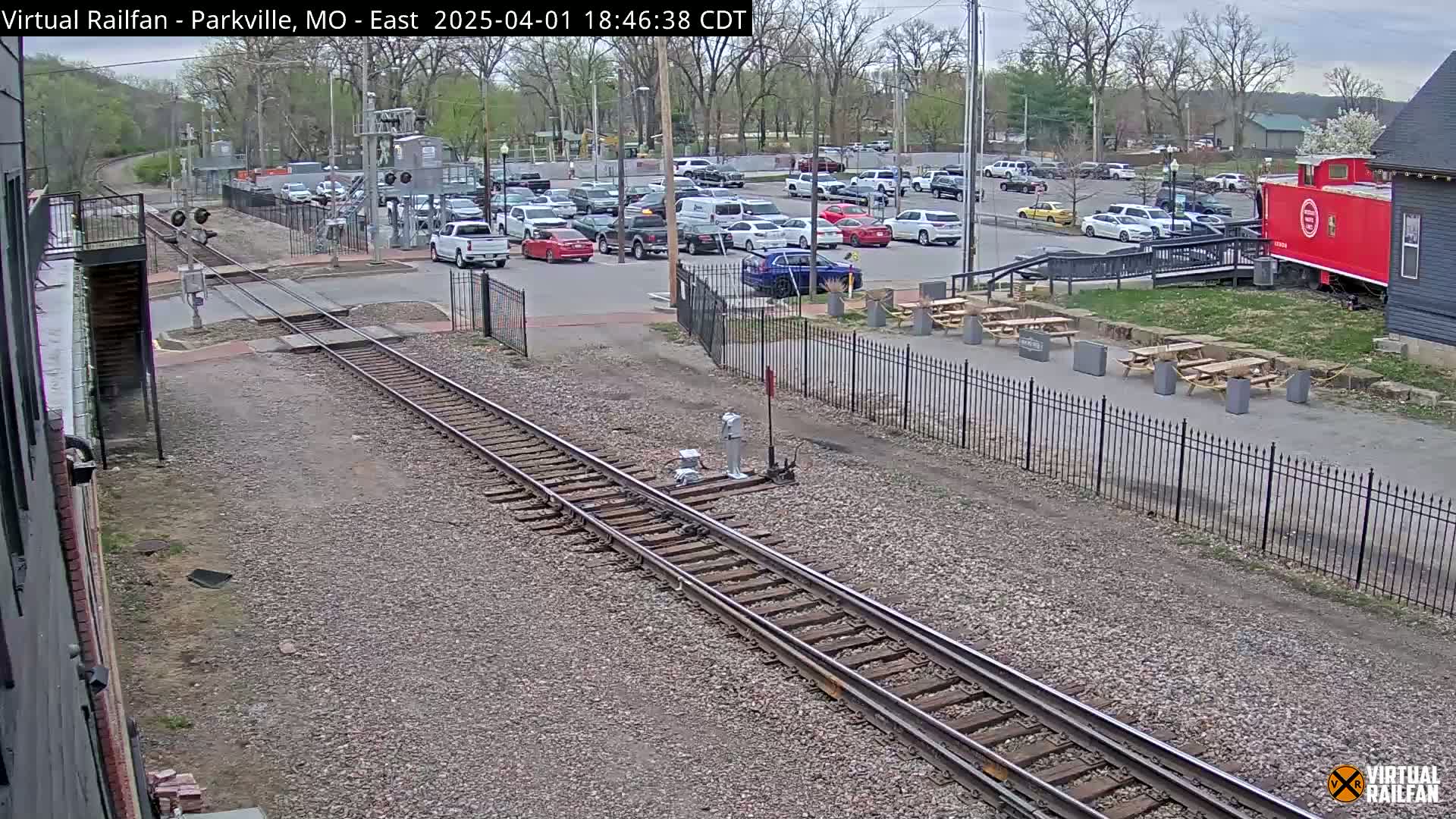A railroad track runs through a parking lot with numerous cars, near a red caboose and picnic tables under a partly cloudy sky.