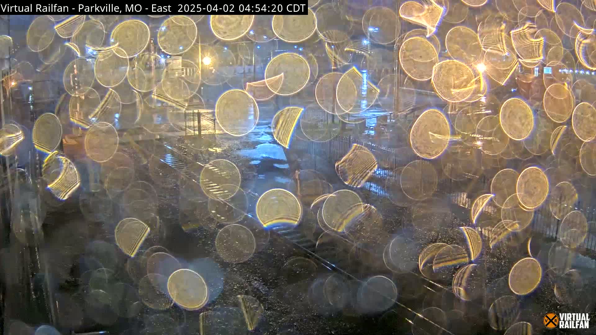 Rain-covered window shows a blurry view of train tracks at night, with lights visible in the background.