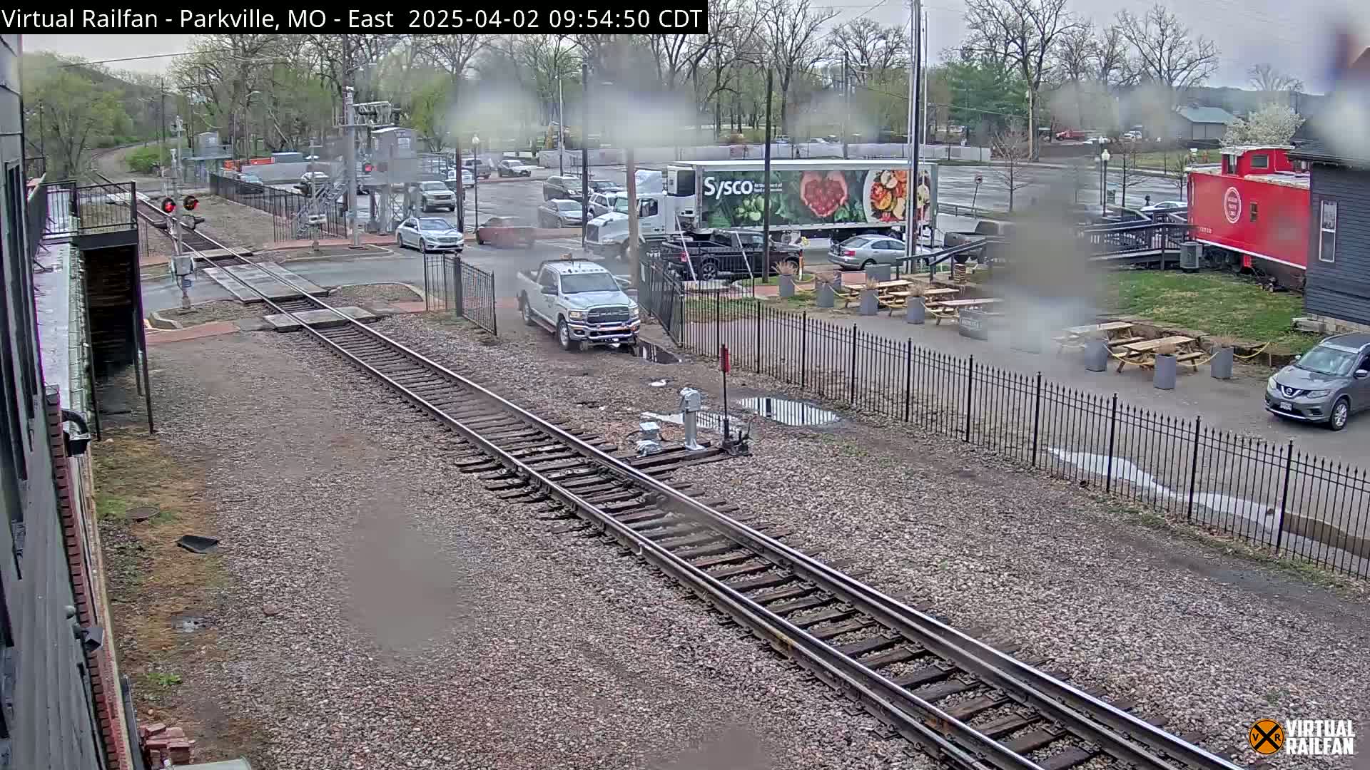 A rainy day view of a railroad crossing with a truck, cars, and a red caboose, near an outdoor seating area.