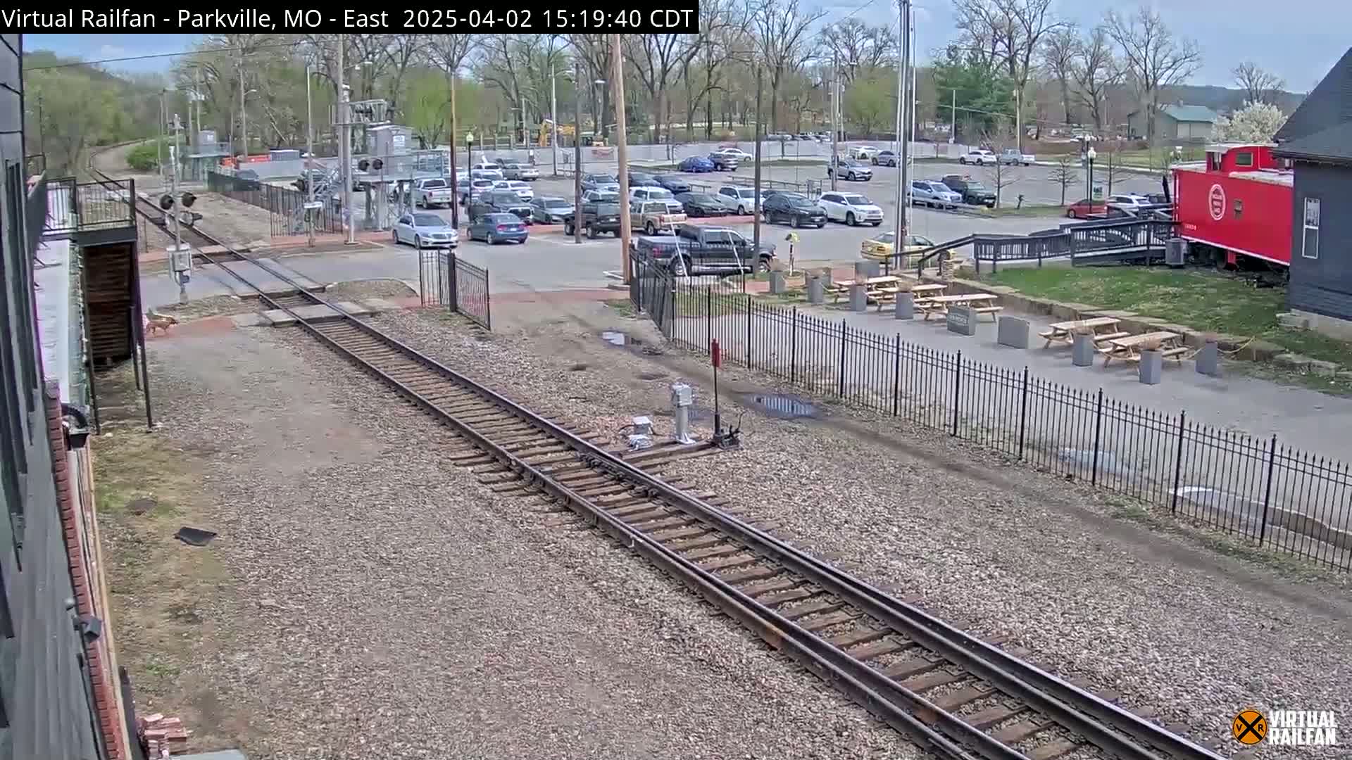 A train track runs beside a parking lot with numerous cars, a red caboose, and outdoor picnic tables under a partly sunny sky.