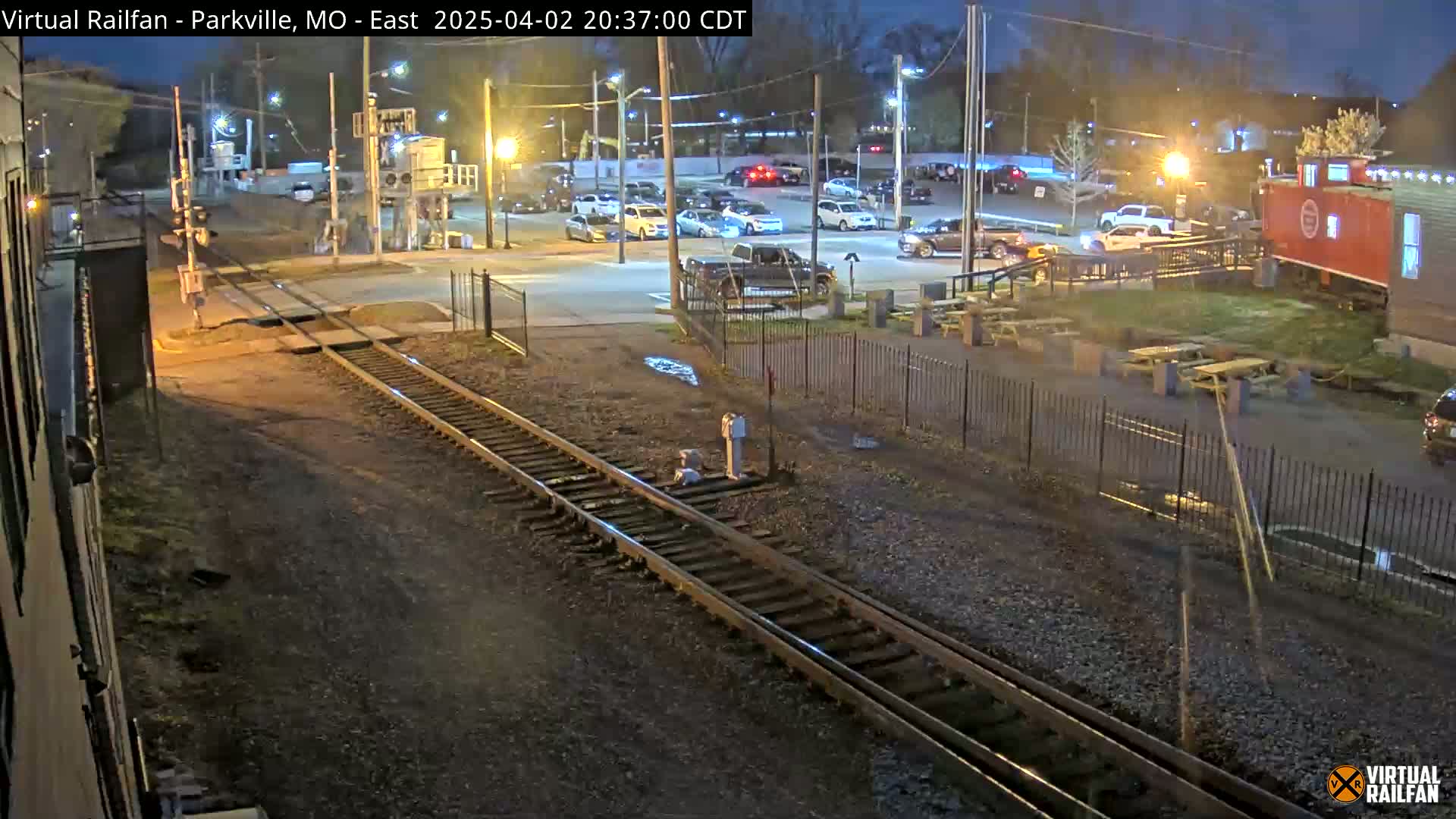 A nighttime view of railroad tracks intersecting a parking lot with several cars and picnic tables, under partly cloudy skies.
