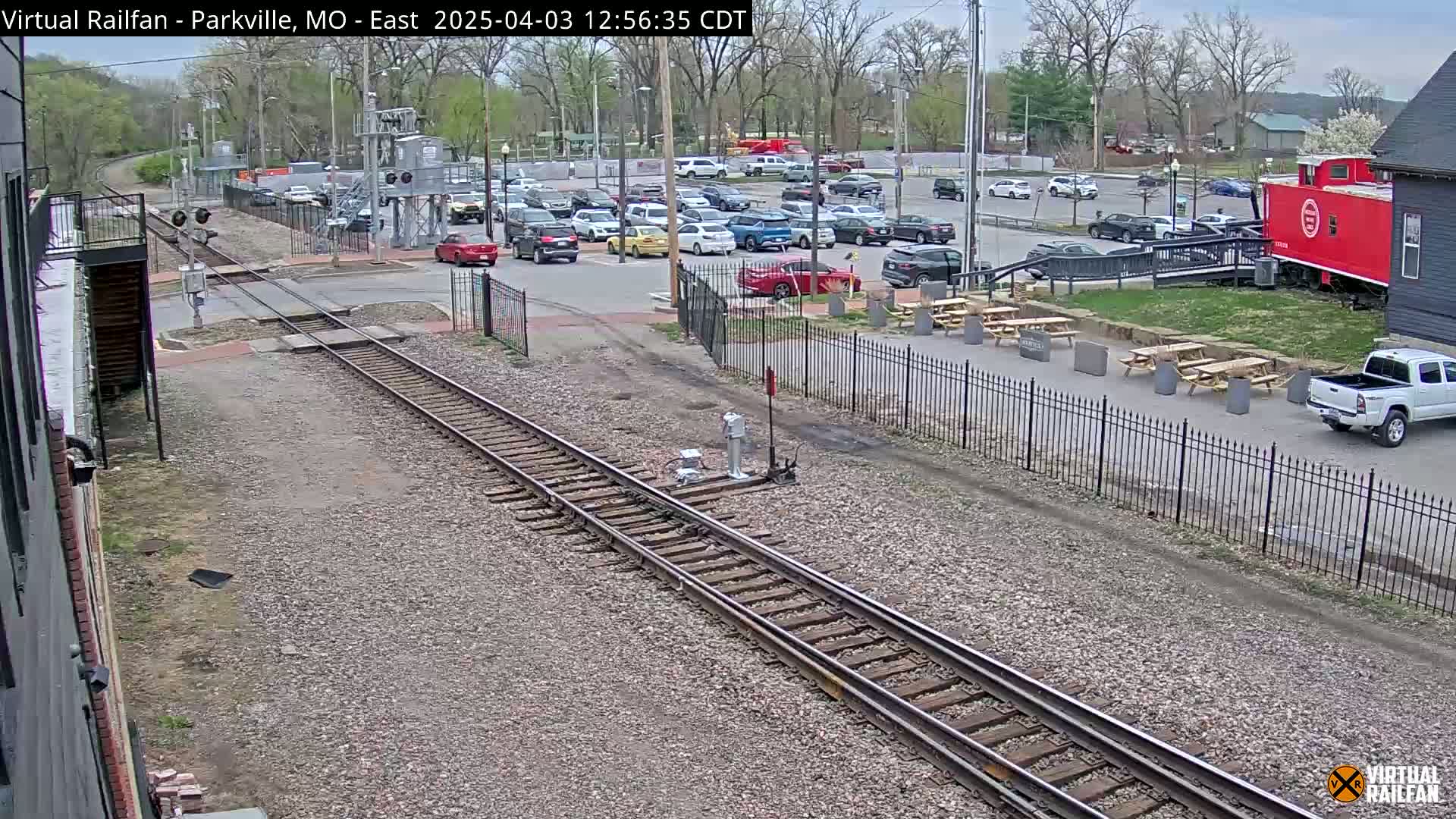 Railroad tracks cross a street next to a parking lot with many cars, picnic tables, and a red caboose under a partly cloudy sky.