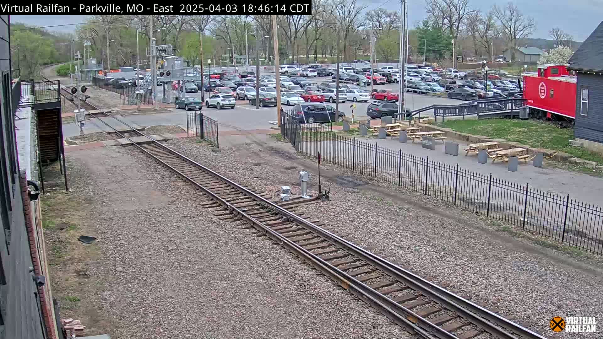 A view of railroad tracks and a large parking lot with cars, next to a building and picnic tables under a partly cloudy sky.