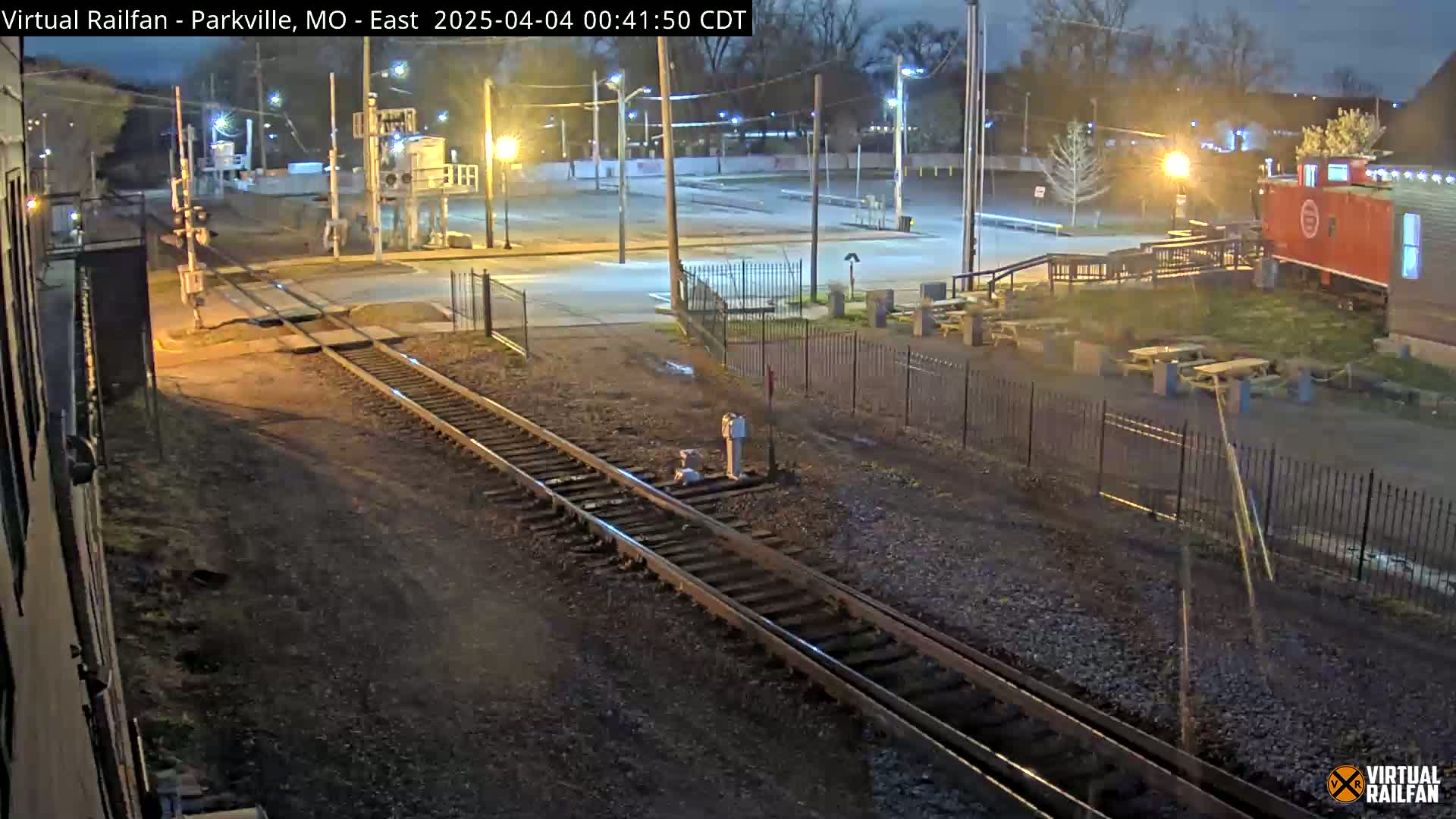 A nighttime view of railroad tracks intersecting with a road, near a fenced-in area with picnic tables and a red caboose-like structure.