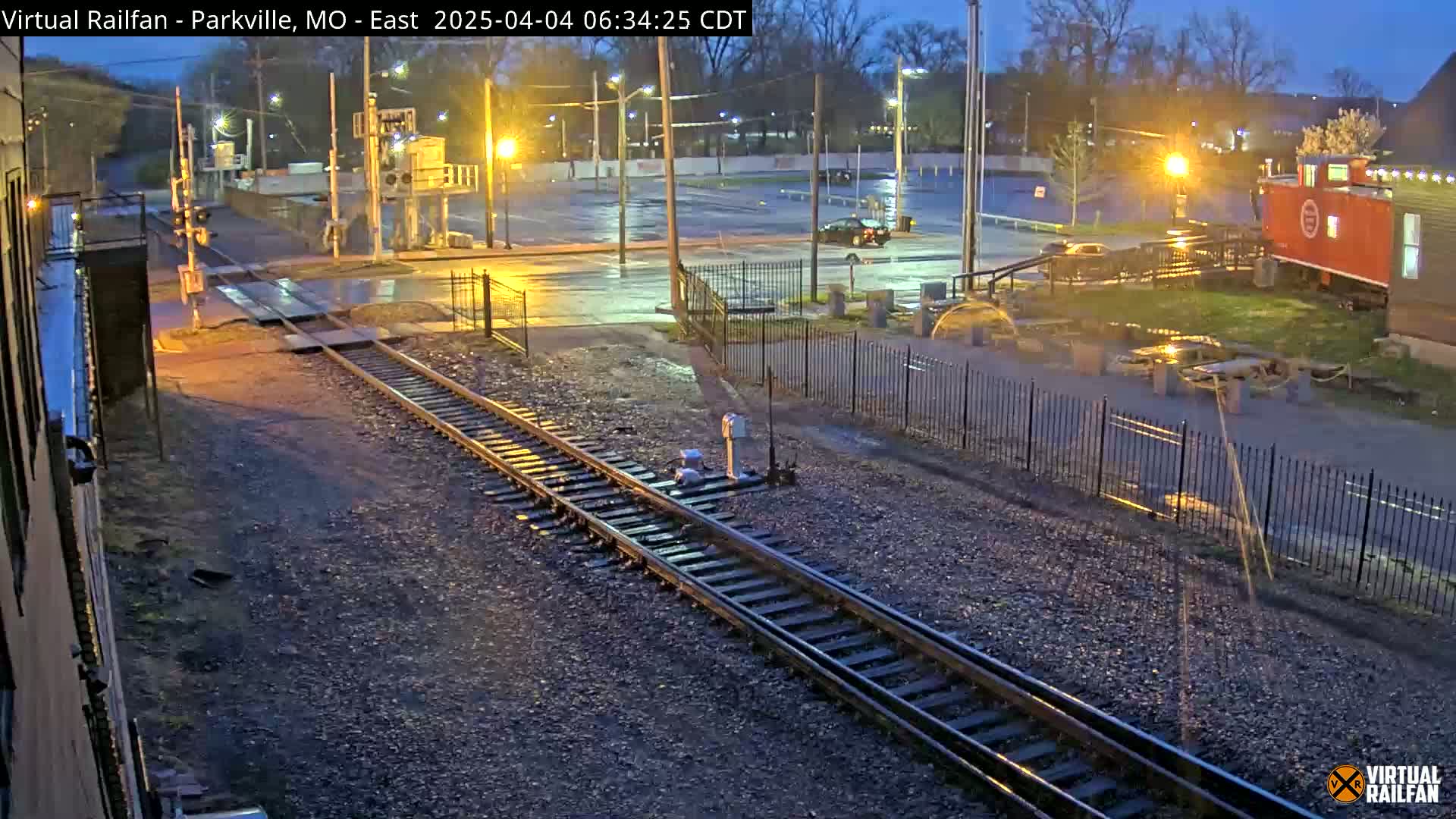 A nighttime view of railroad tracks intersecting with a street, featuring a mostly empty parking lot and a red caboose in the background, under dark, wet conditions.