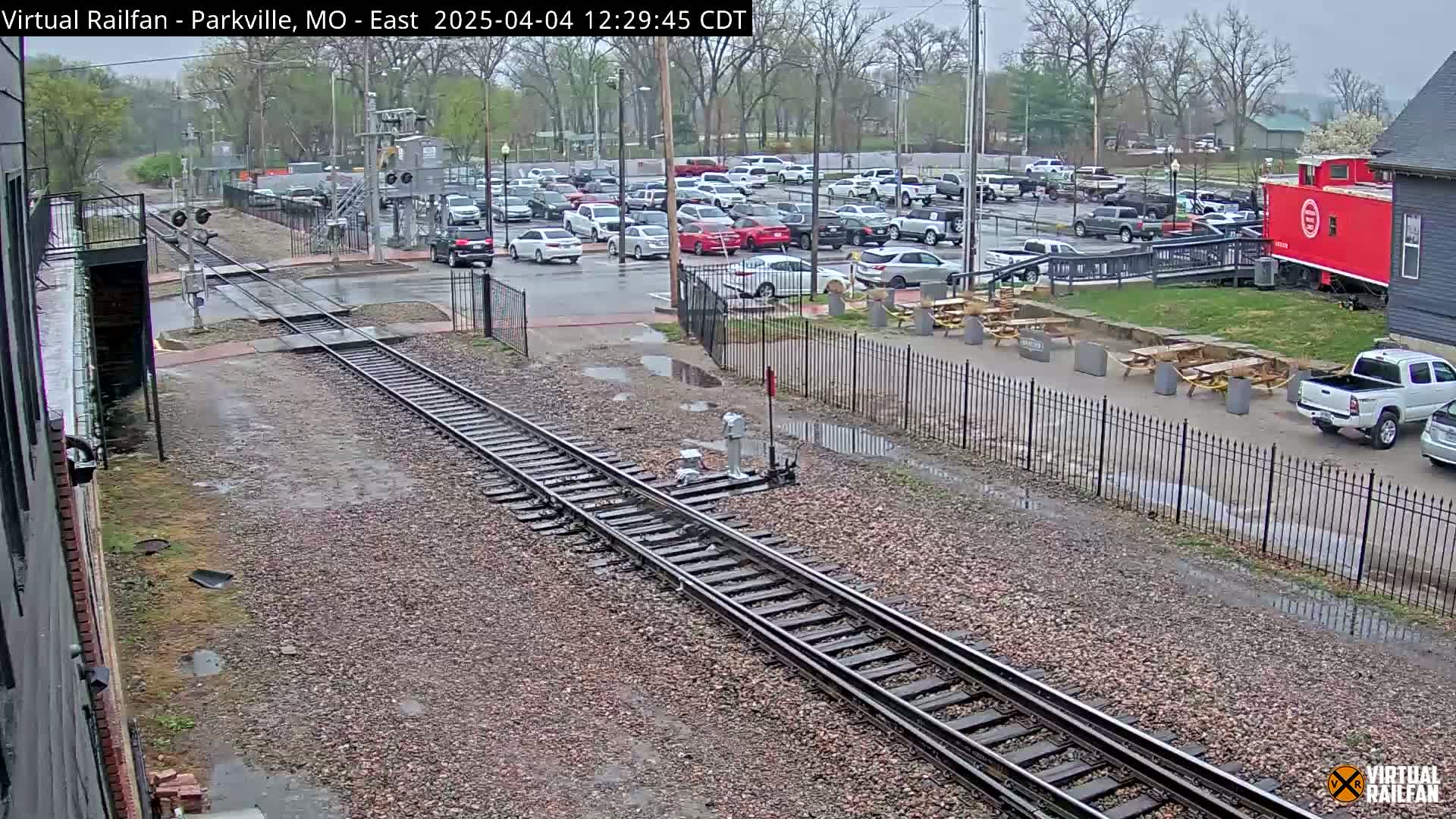 Railroad tracks in a parking lot on a rainy day, next to a red caboose and several picnic tables.