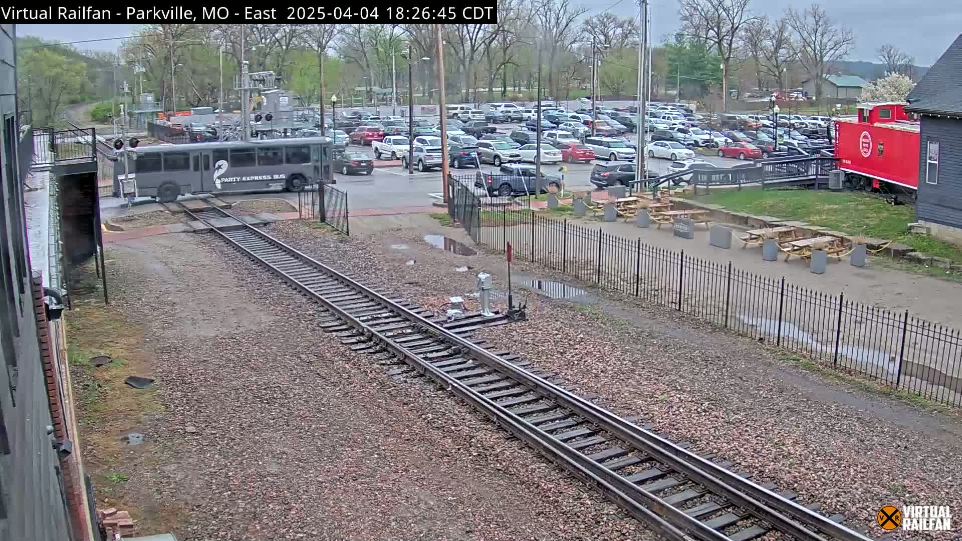 A gray bus is crossing railroad tracks near a parking lot and red caboose under an overcast sky.