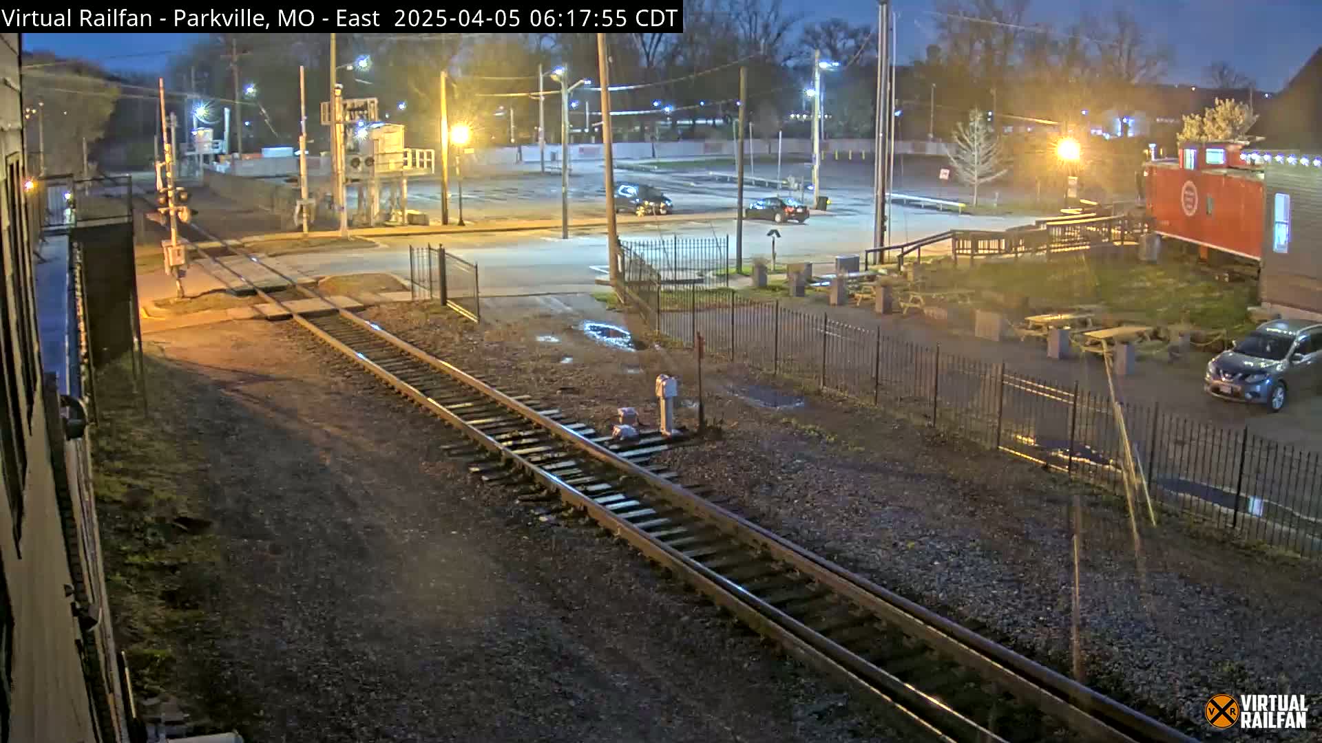 A nighttime view of railroad tracks intersecting with a road near a parking lot and a building, with several cars present and puddles in the ground.