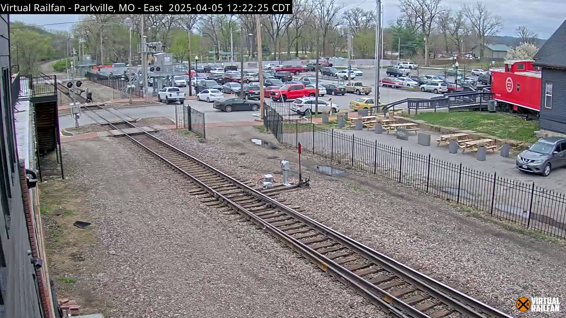 A railroad track runs through a parking lot with several cars, picnic tables, and a red caboose under a partly cloudy sky.