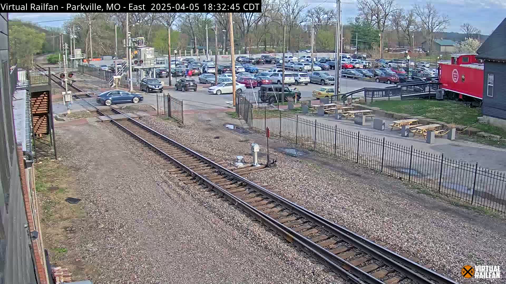 A mostly sunny day shows a railroad crossing with a car passing over the tracks near a parking lot full of cars and a red caboose next to a building.