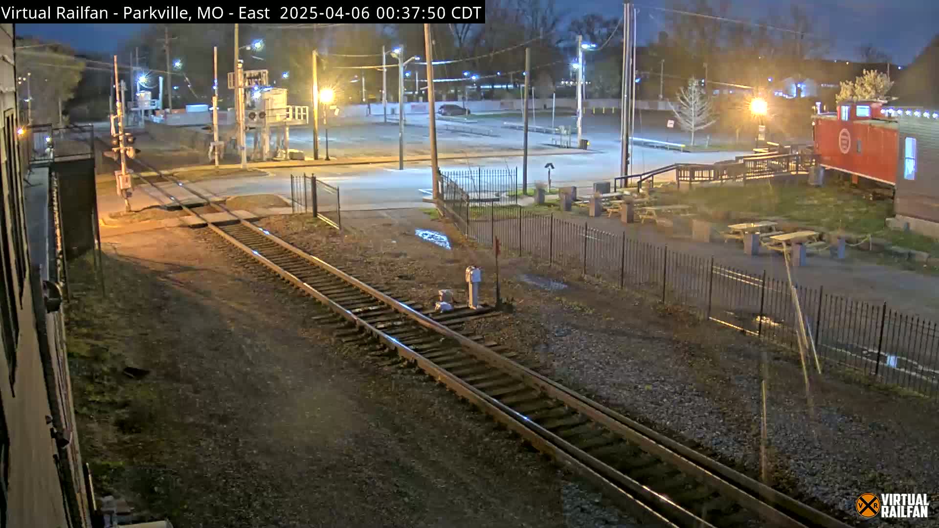 A nighttime view of railroad tracks intersecting a road, with a mostly empty parking lot and a red caboose-like structure visible in the background.