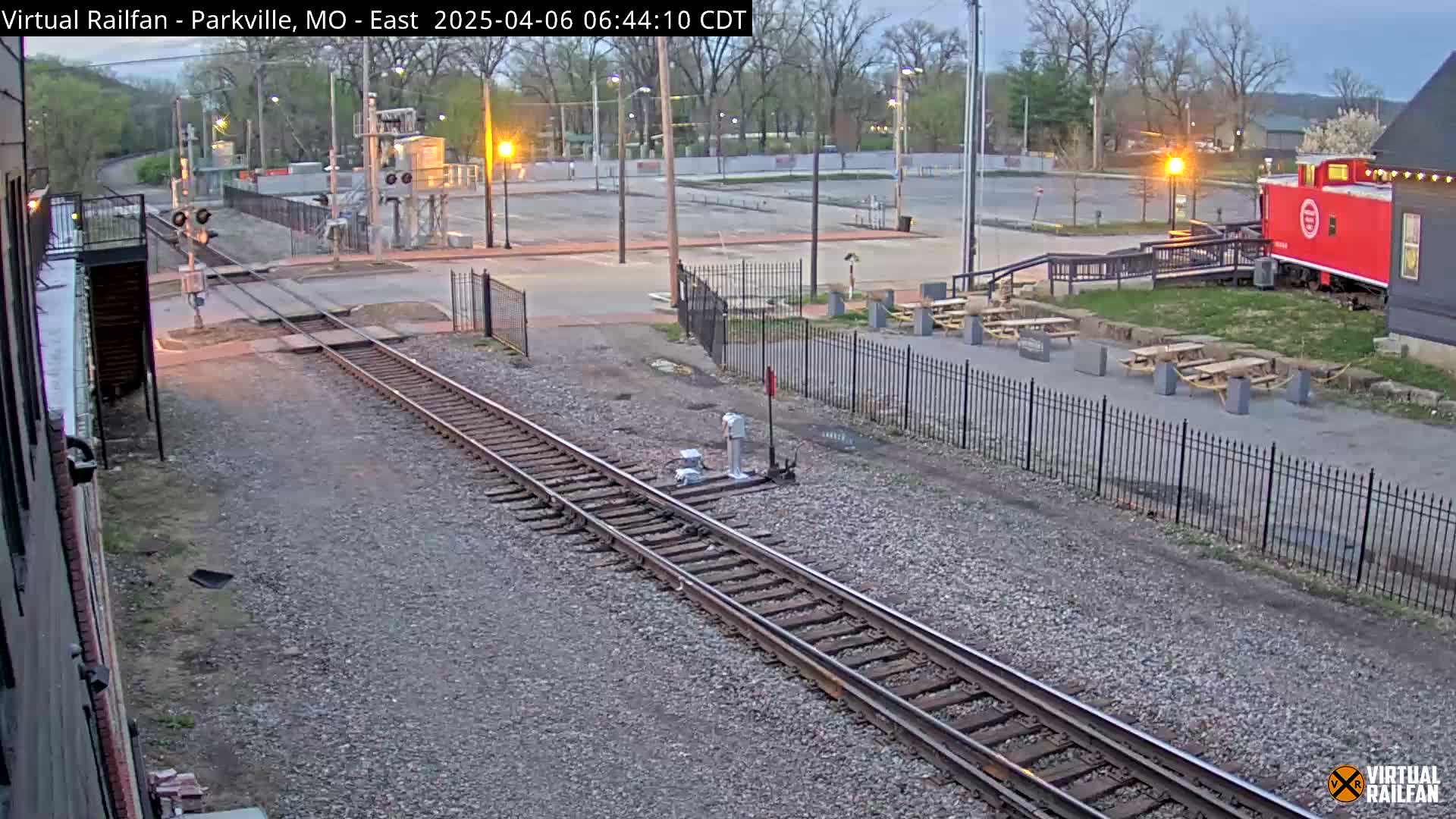 Railroad tracks extend from a train crossing near a parking lot with picnic tables and a red caboose under a twilight sky.