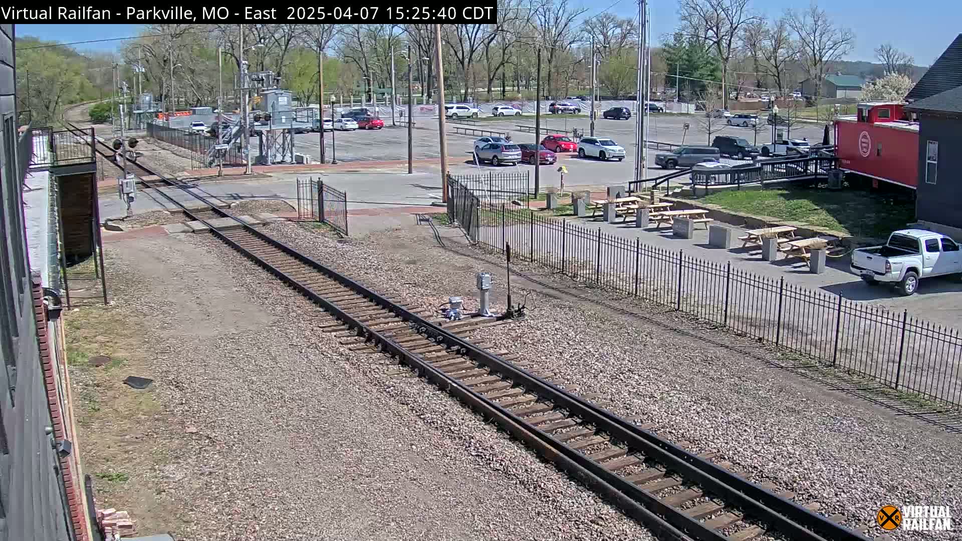 Railroad tracks run alongside a parking lot with cars and picnic tables under a sunny sky.