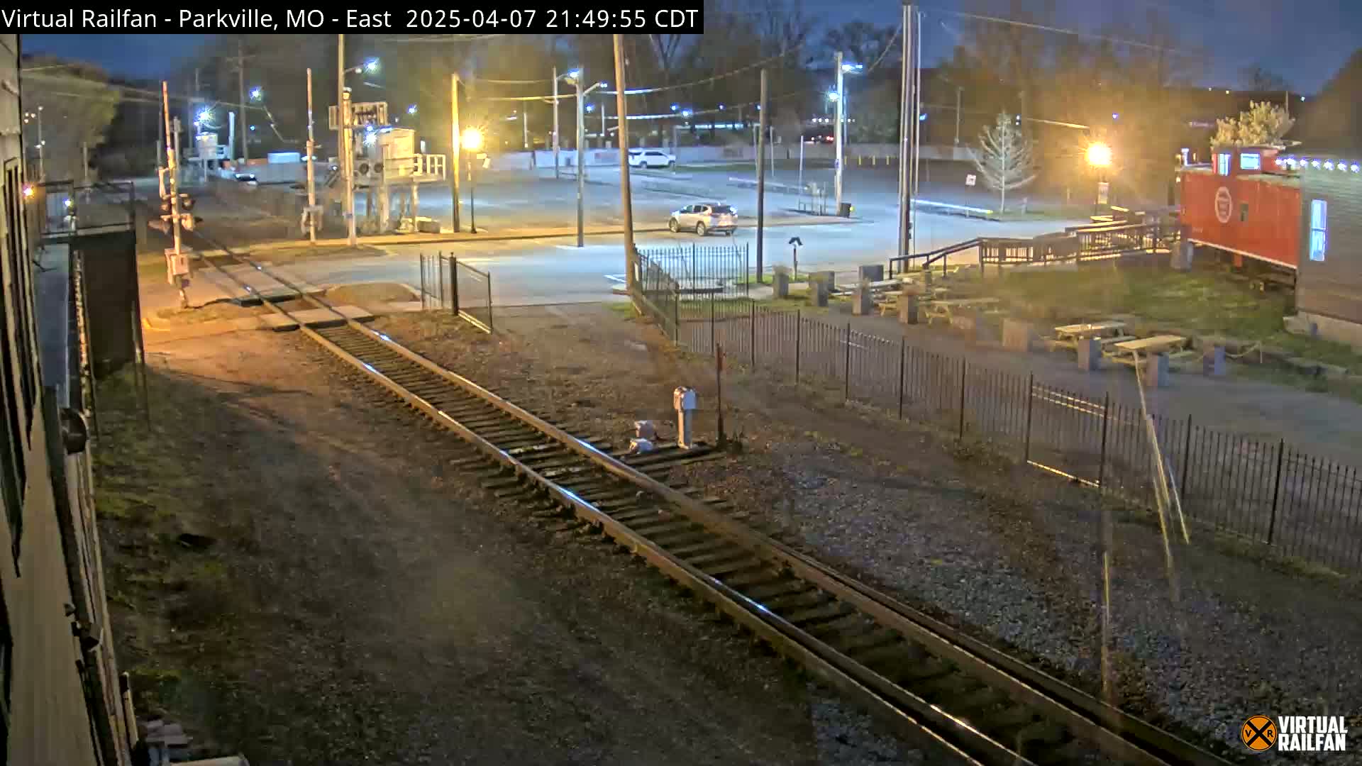 A nighttime view of railroad tracks intersecting a road near a parking lot with a few cars and picnic tables next to a red building.
