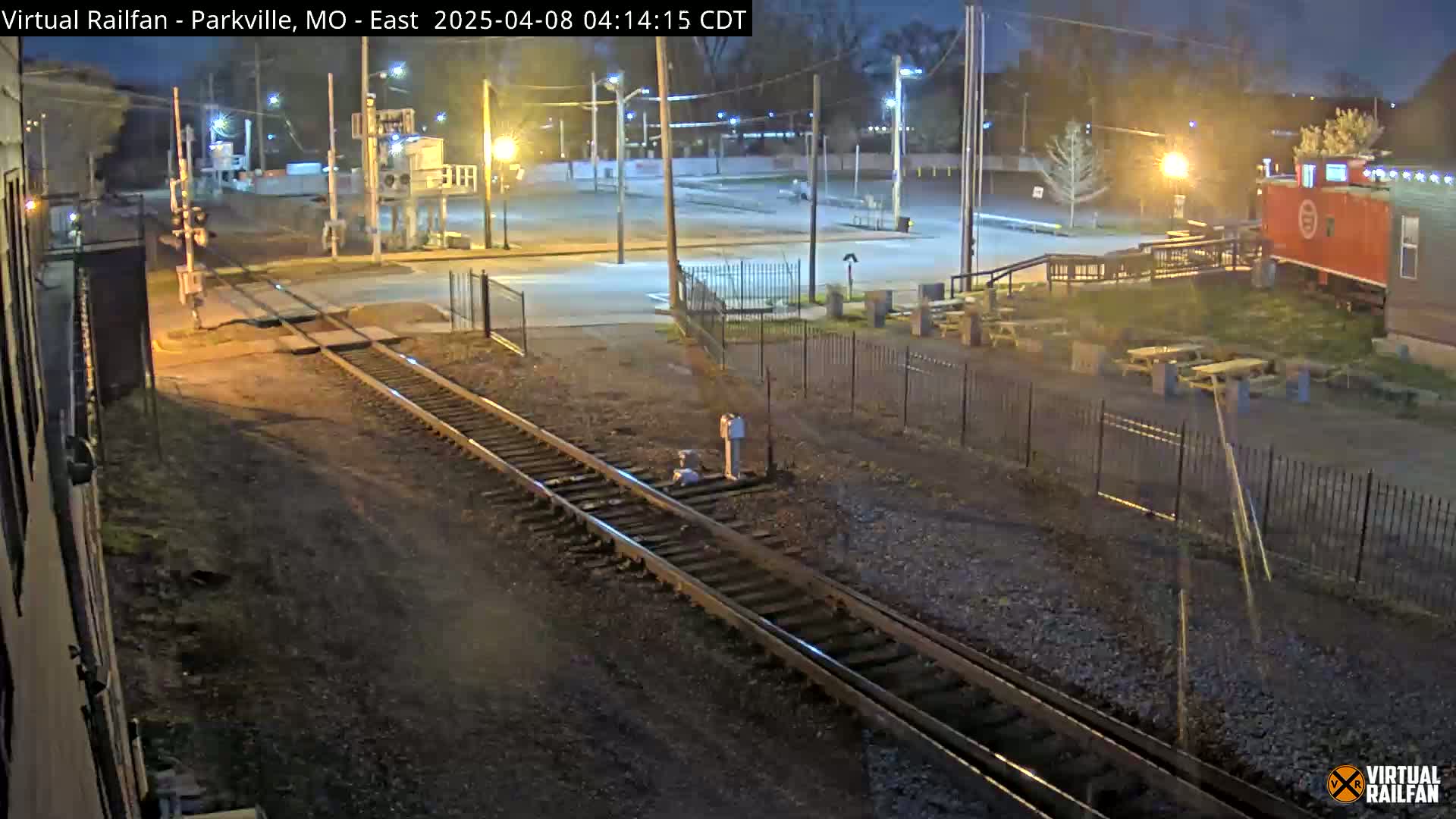A nighttime view of railroad tracks intersecting a road near a fenced-in area with picnic tables and a red building, under dimly lit conditions.