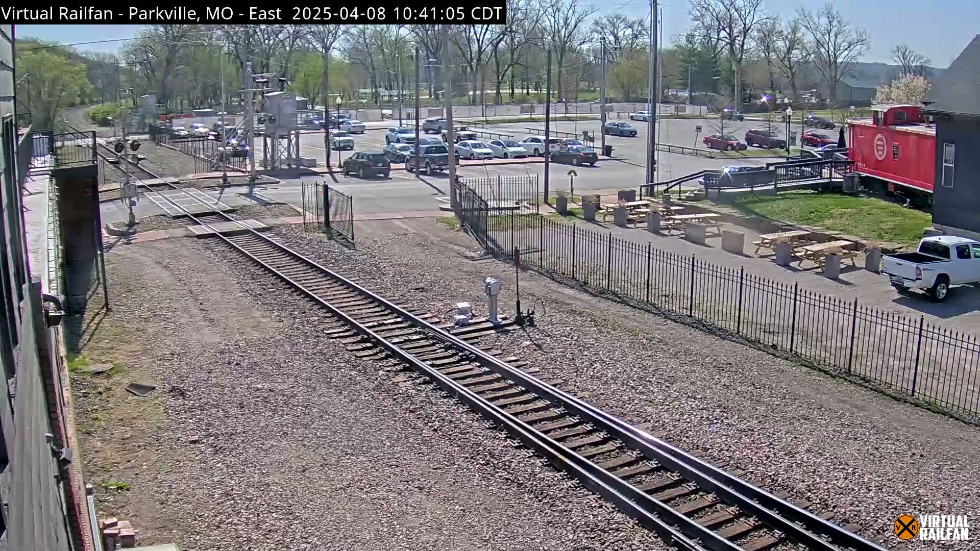 A sunny outdoor scene shows railroad tracks intersecting with a road near a parking lot and picnic area with a red caboose visible in the background.