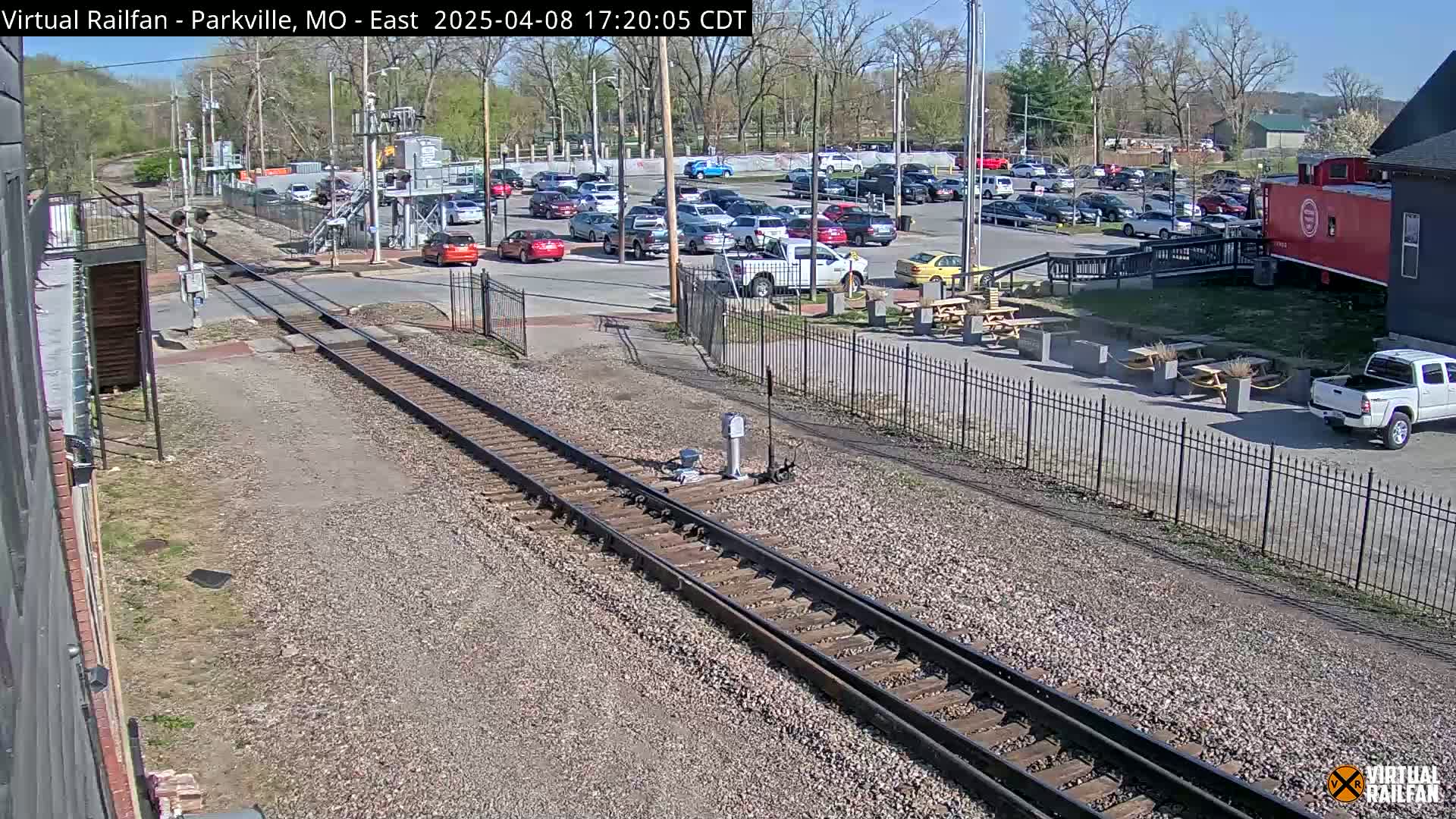 A sunny day view of railroad tracks intersecting with a road next to a parking lot, a building with outdoor seating, and a red caboose.