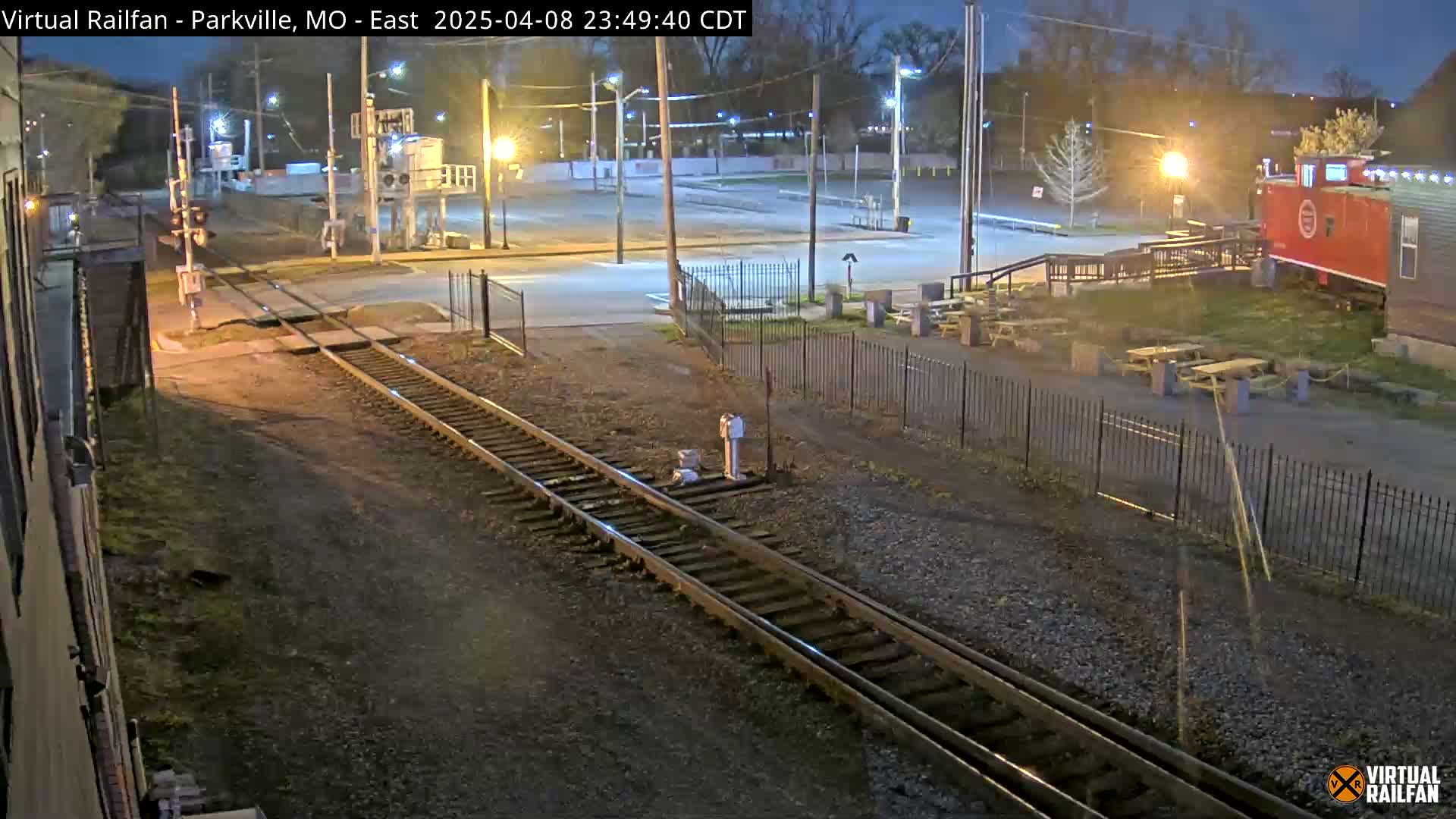 A nighttime view of railroad tracks intersecting a street near a fenced-in area with picnic tables and a red caboose.
