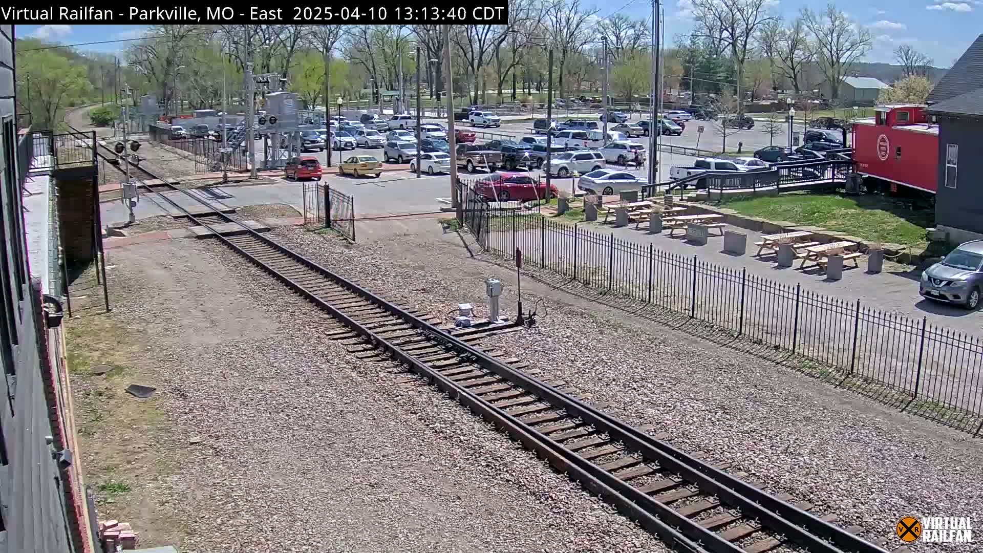 A sunny day shows a railroad crossing with a parking lot full of cars, picnic tables, and a red caboose next to a building.