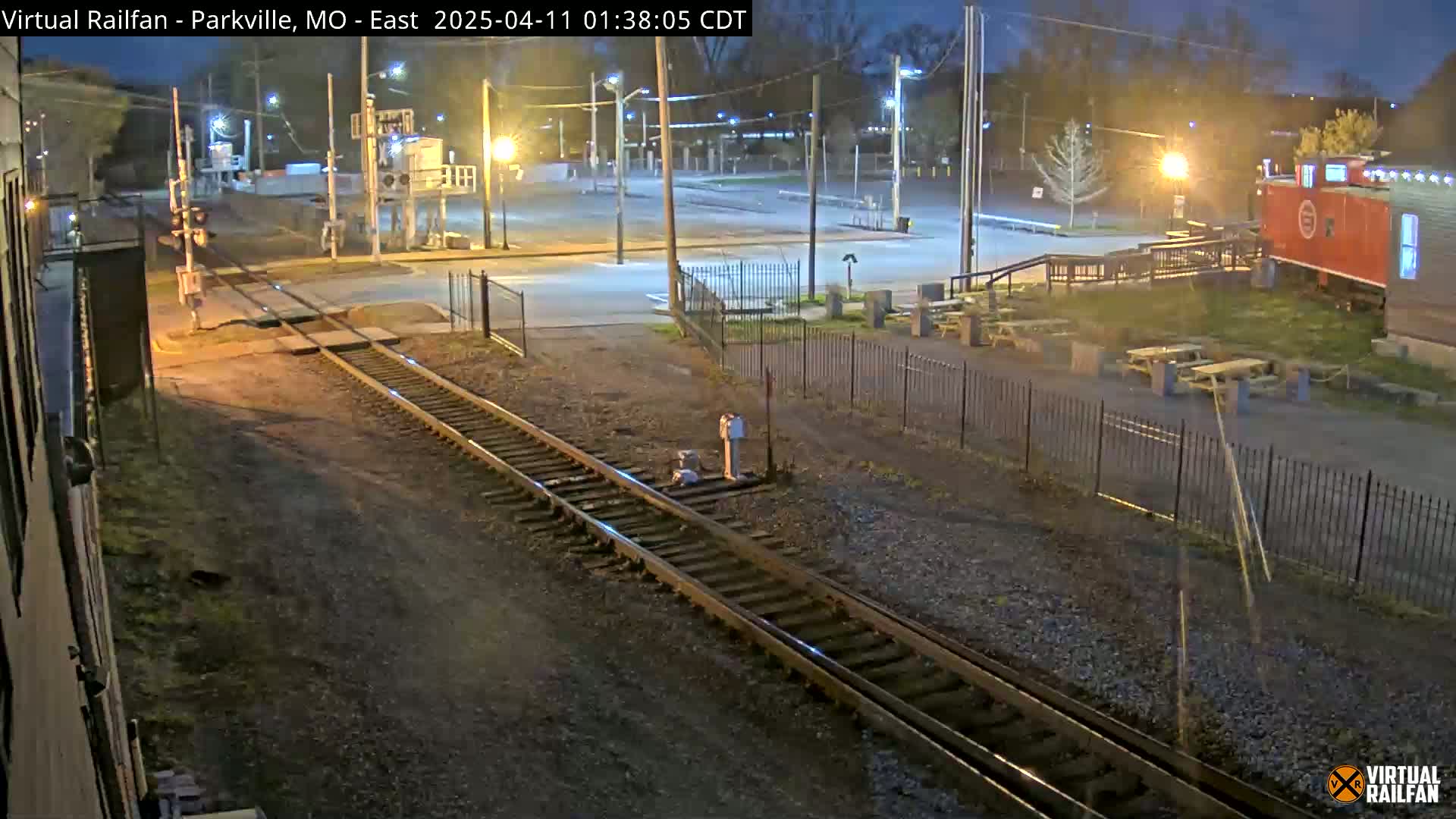 A nighttime view of railroad tracks intersecting a road, with a parking lot, some buildings, and picnic tables visible under artificial lighting.