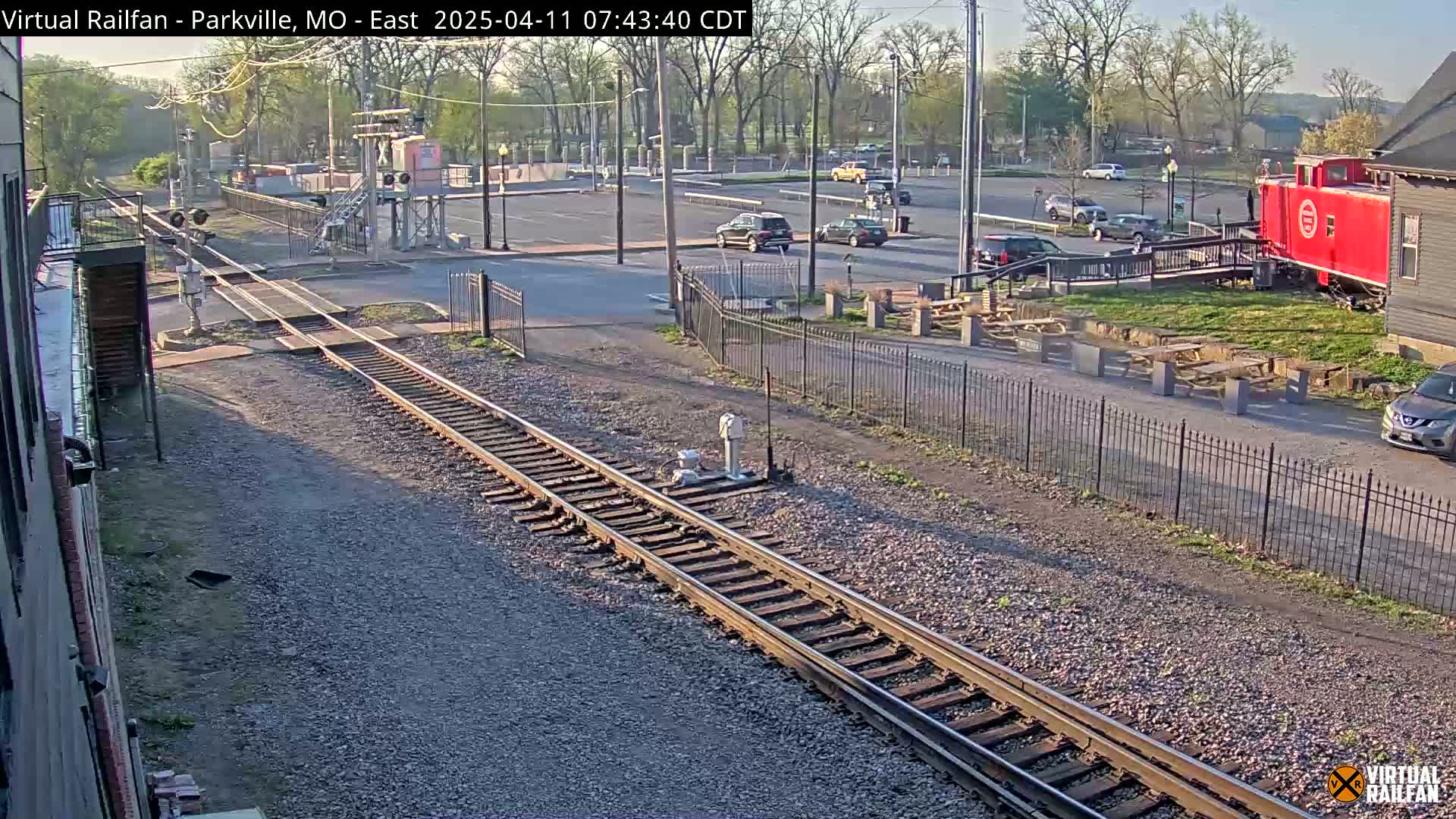 A sunny view of railroad tracks, a red caboose, a parking lot with several cars, and outdoor seating areas.