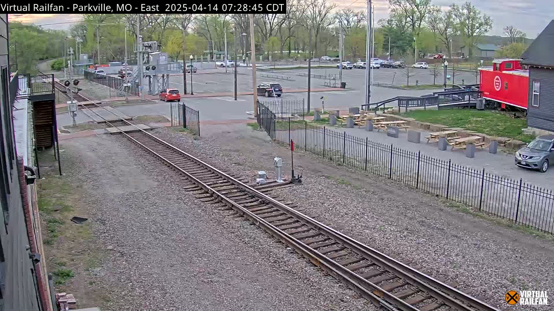 A view of railroad tracks next to a parking lot with several cars, picnic tables, and a red caboose under an overcast sky.